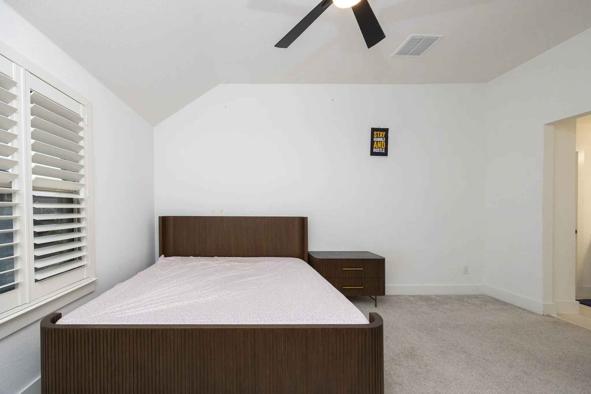 White plantation shutters, sloped ceiling, overhead ceiling fan, and light gray carpeting