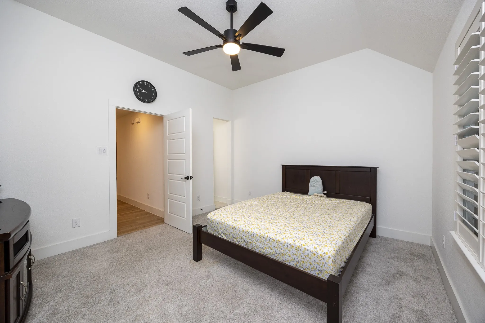 Vaulted ceiling with a ceiling fan, plush gray carpeting, white plantation shutters, and a paneled interior door with matte black hardware