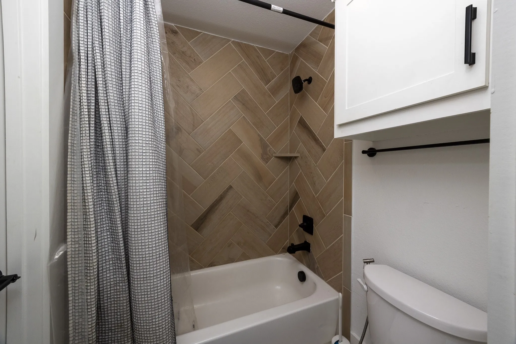 Bathtub with herringbone tile surround, matte black fixtures, built-in shower niche, and white wall-mounted cabinetry