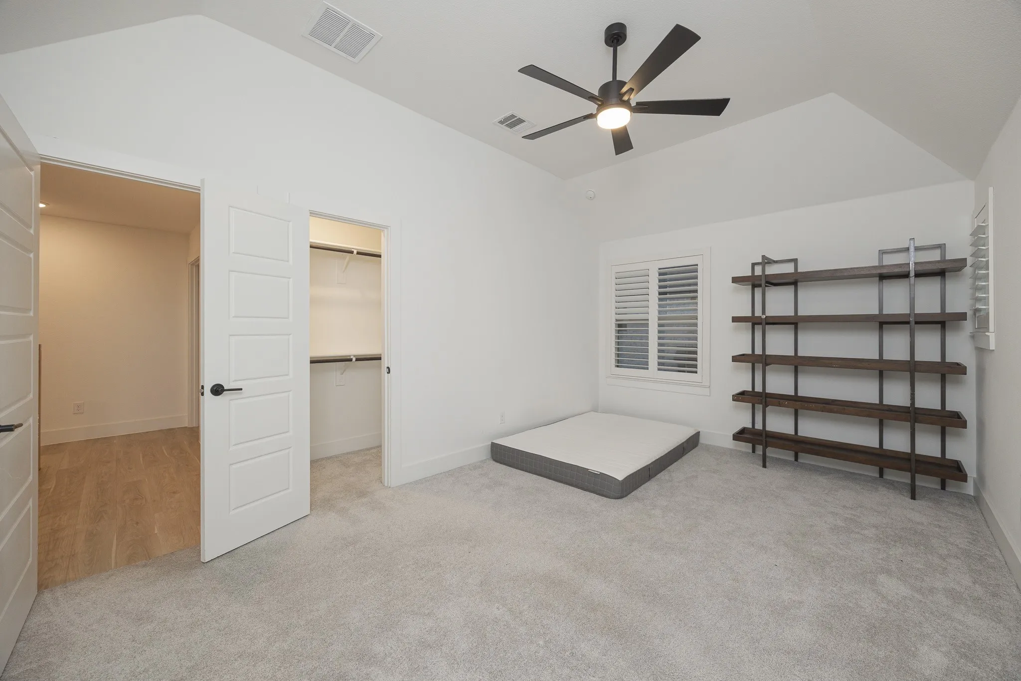 Vaulted ceiling with an attached ceiling fan, light-toned carpeting, window with plantation shutters, and a built-in closet with shelving