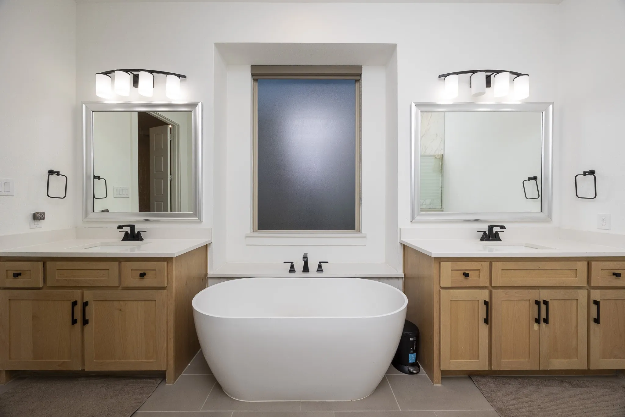 Freestanding bathtub with a black wall-mounted faucet, dual vanities with light wood-finish cabinetry, white countertops, and framed mirrors
