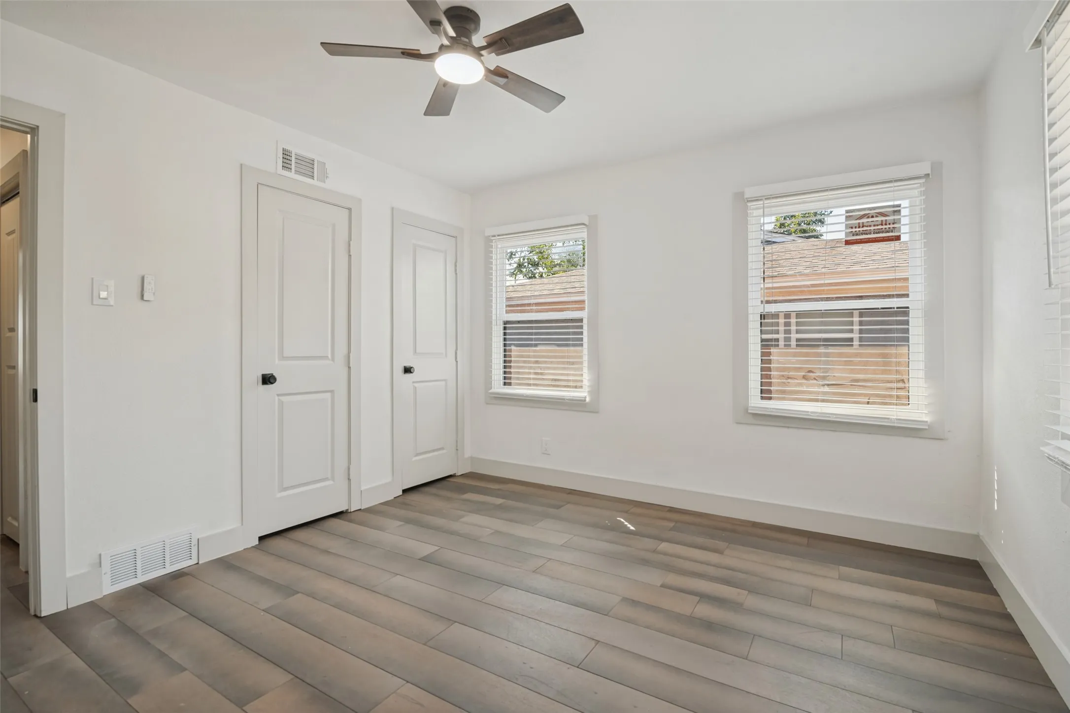 Wood-finish flooring, white walls, two paneled interior doors, two windows, and a ceiling fan with integrated lighting
