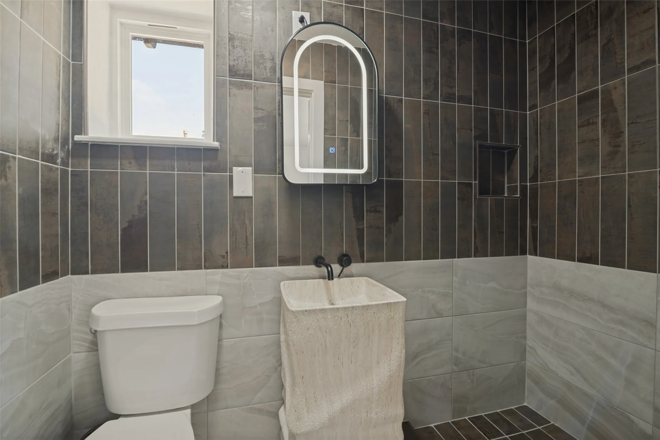 Floor-to-ceiling tilework, modern pedestal sink, illuminated arched mirror, and a tiled shower​​‌​​​​‌​​‌‌​‌‌​​​‌‌​‌​‌​‌​​​‌​​ niche
