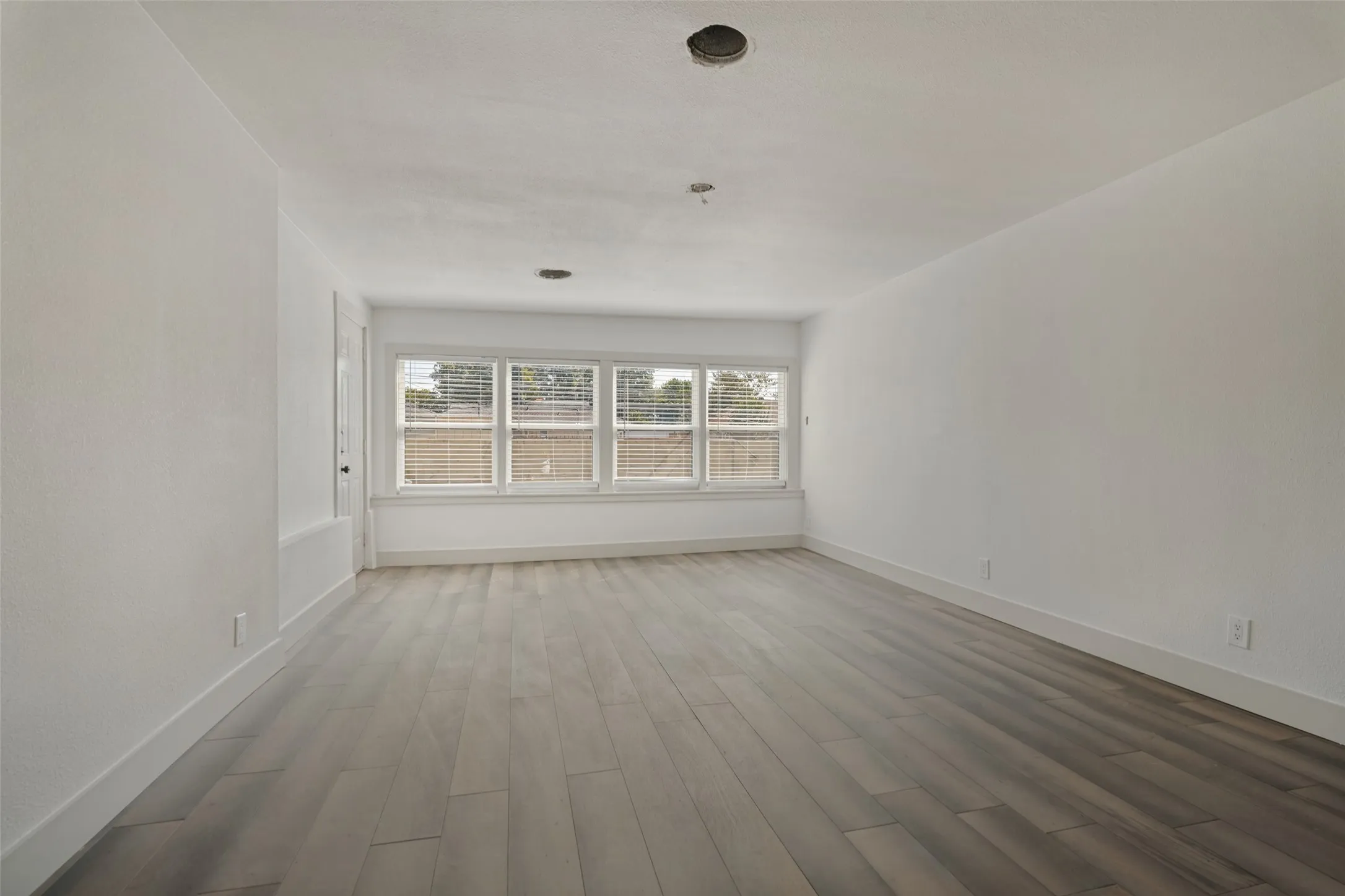 Spacious room featuring wood-finish flooring, white walls, and multiple windows with blinds