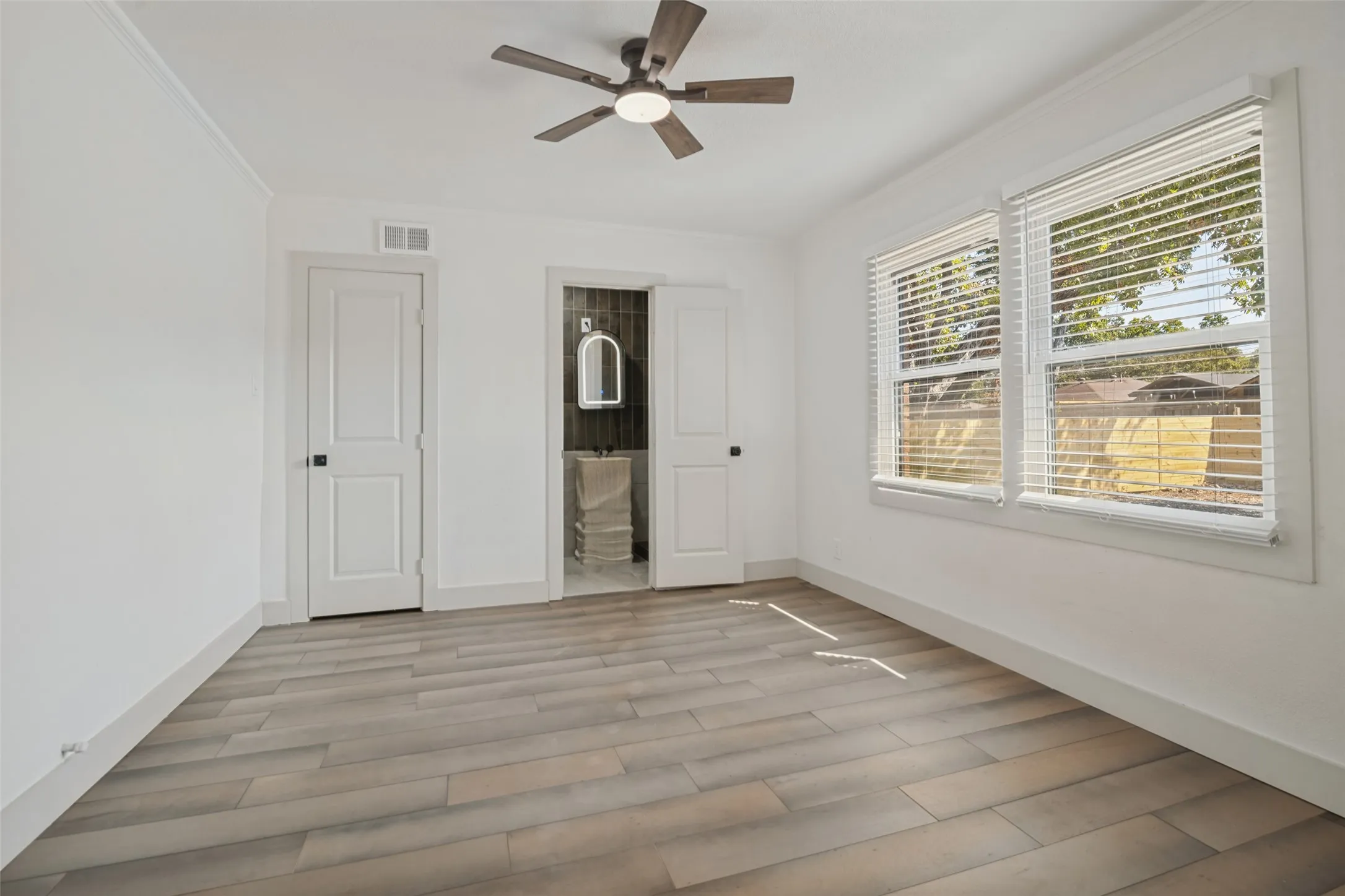 Spacious room featuring wood-finish flooring, two windows with blinds, a ceiling fan, and access to an adjoining bathroom with a pedestal sink and mirror