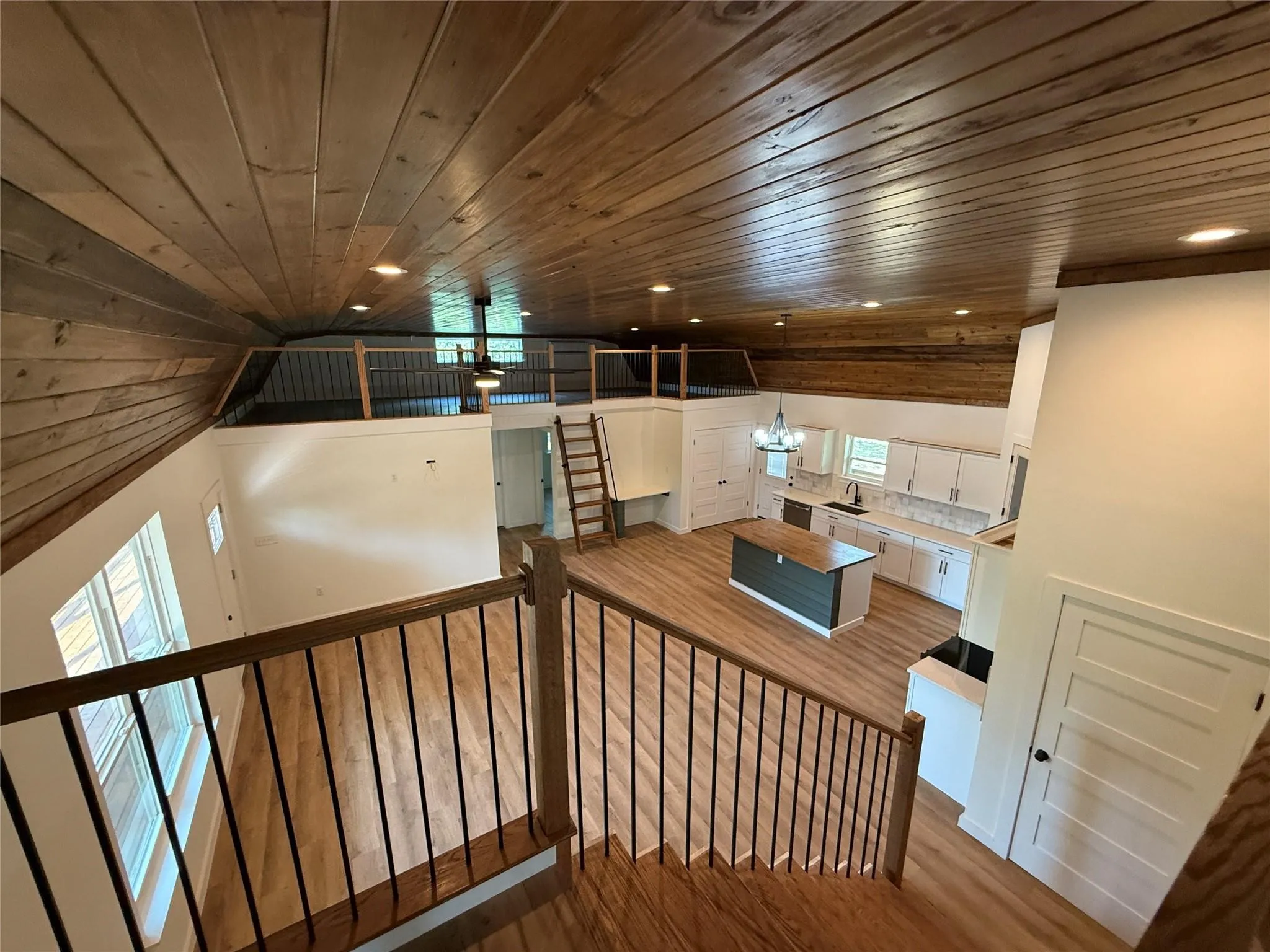 Wood-finish flooring throughout, exposed wood-paneled ceiling, white shaker cabinetry, kitchen island with wood countertop, and black metal railing