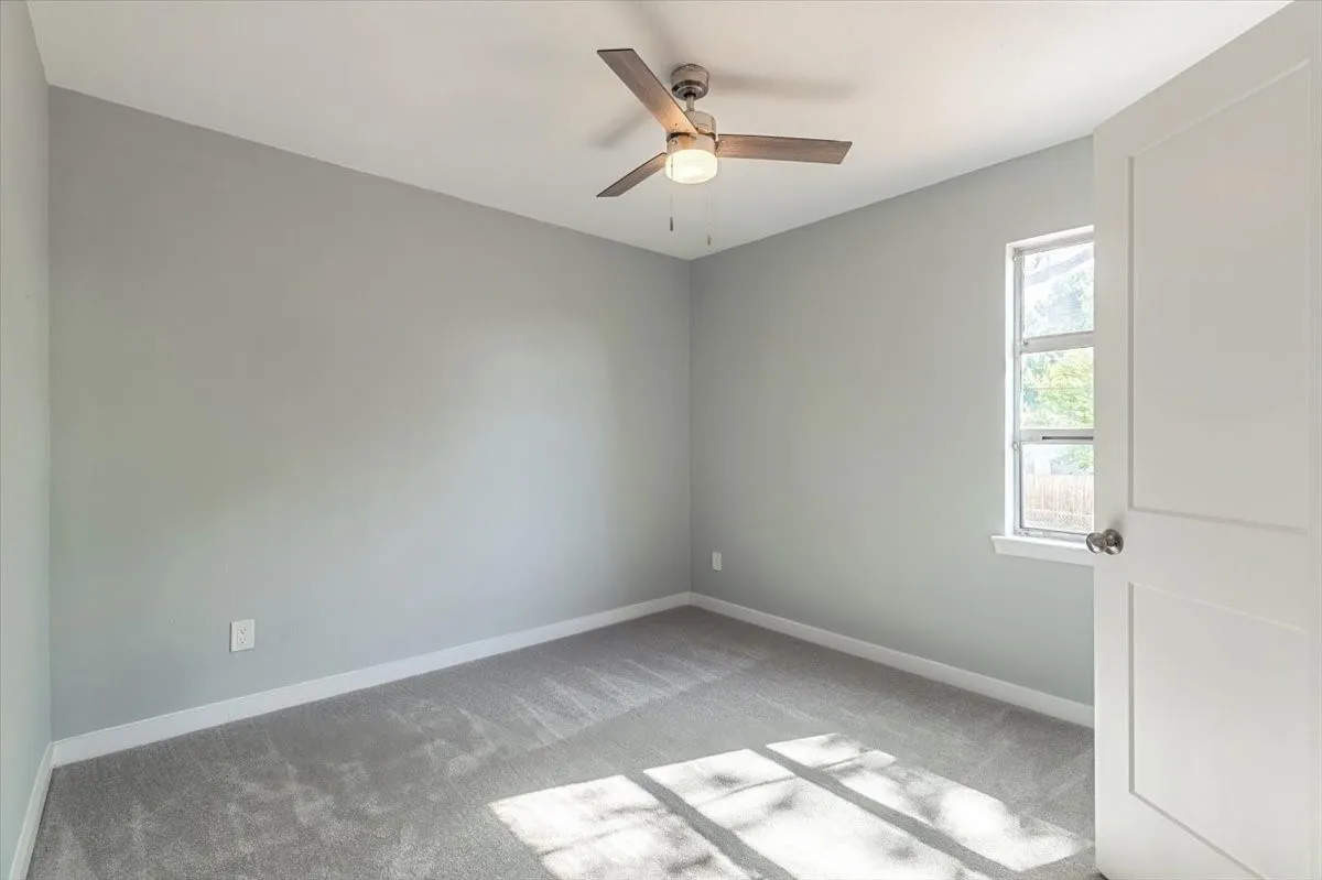 Neutral gray wall paint with white trim, gray carpeting, a single window, and a ceiling fan with integrated lighting