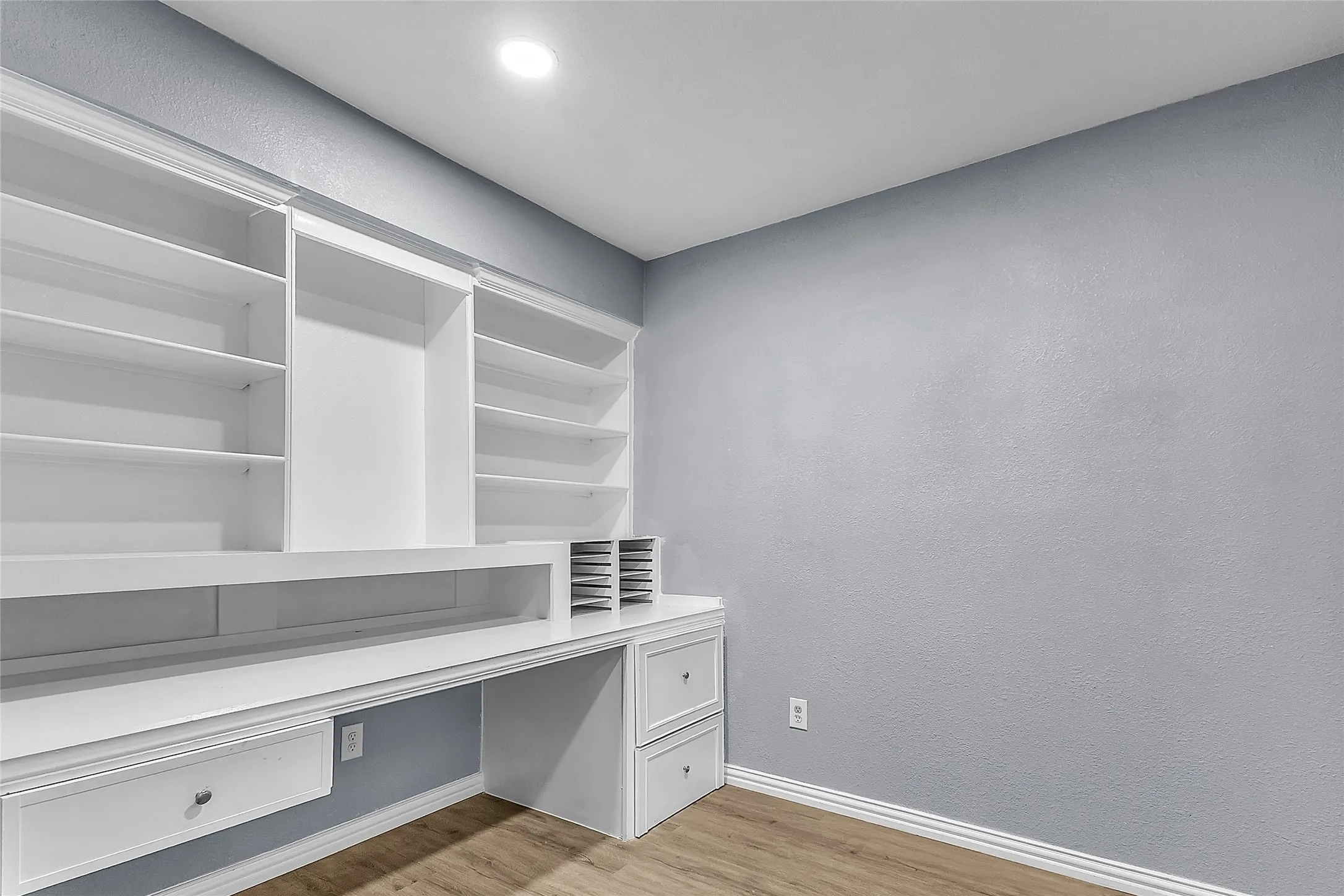 Home office with white built-in shelving and desk against a light gray wall, featuring recessed lighting and a laminate wood floor.