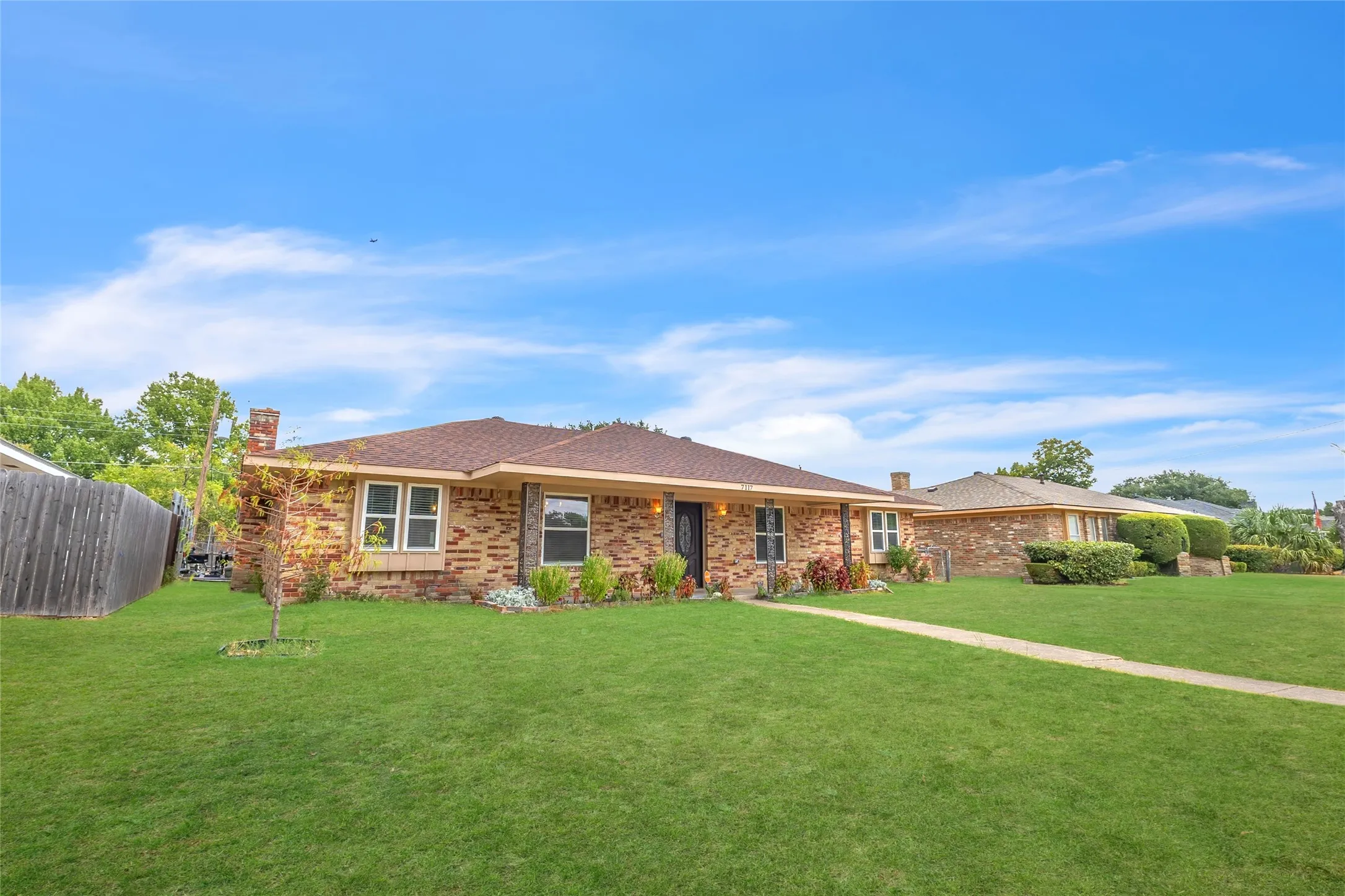 Single-story brick home with a sloped roof, front porch, landscaped yard, and an open layout, bordered by neighboring homes and clear blue sky.