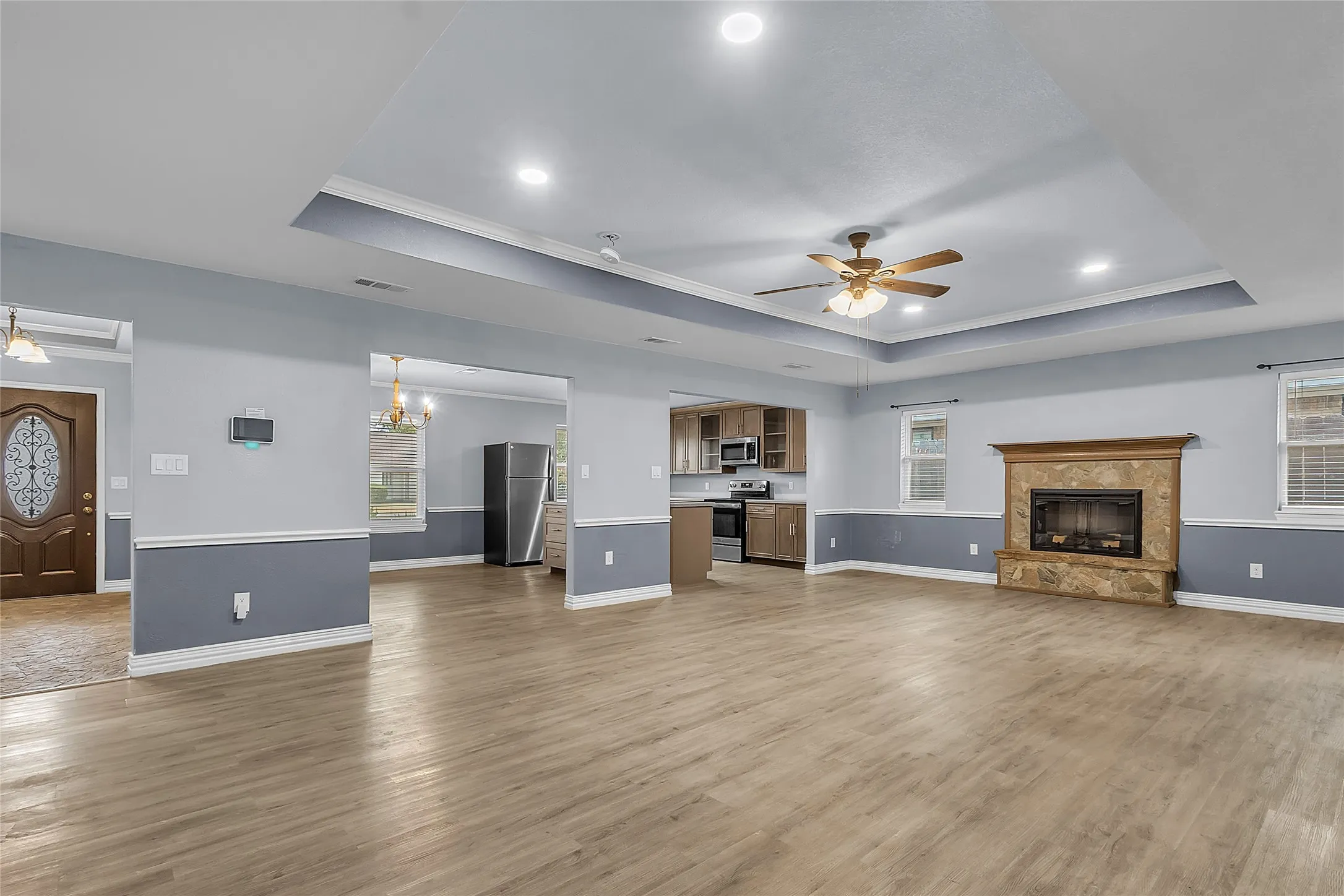 Open-plan living area with hardwood flooring, recessed lighting, and a stone fireplace, accented by a tray ceiling and warm natural light from windows.