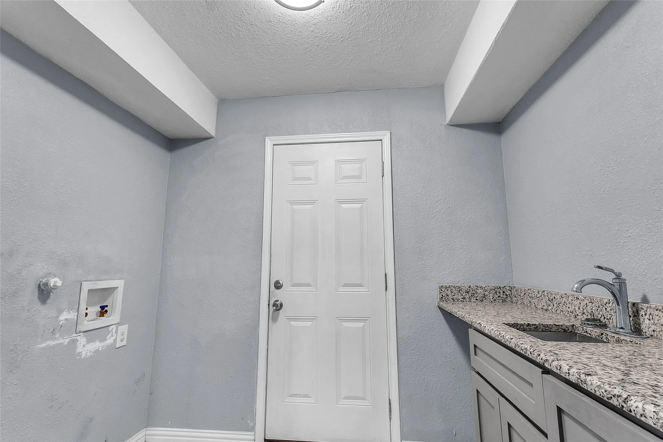 Laundry room with granite countertop, stainless steel sink, and a door leading to an exterior entrance. Textured gray walls enhance the bright atmosphere.