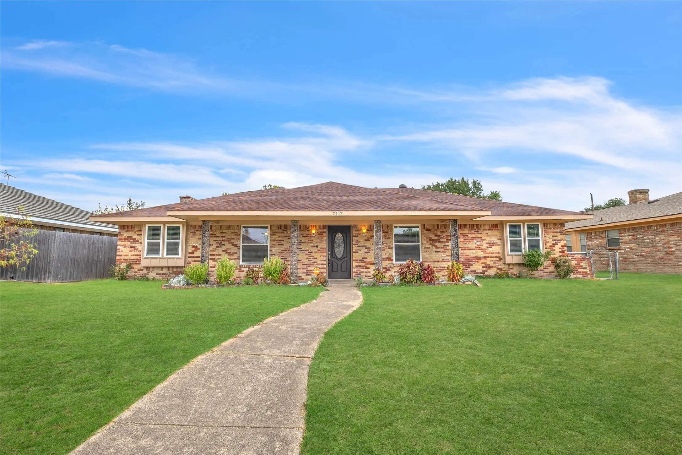 Single-story brick home with a landscaped front yard, a concrete pathway, and a symmetrical facade under a brown shingle roof against a blue sky
