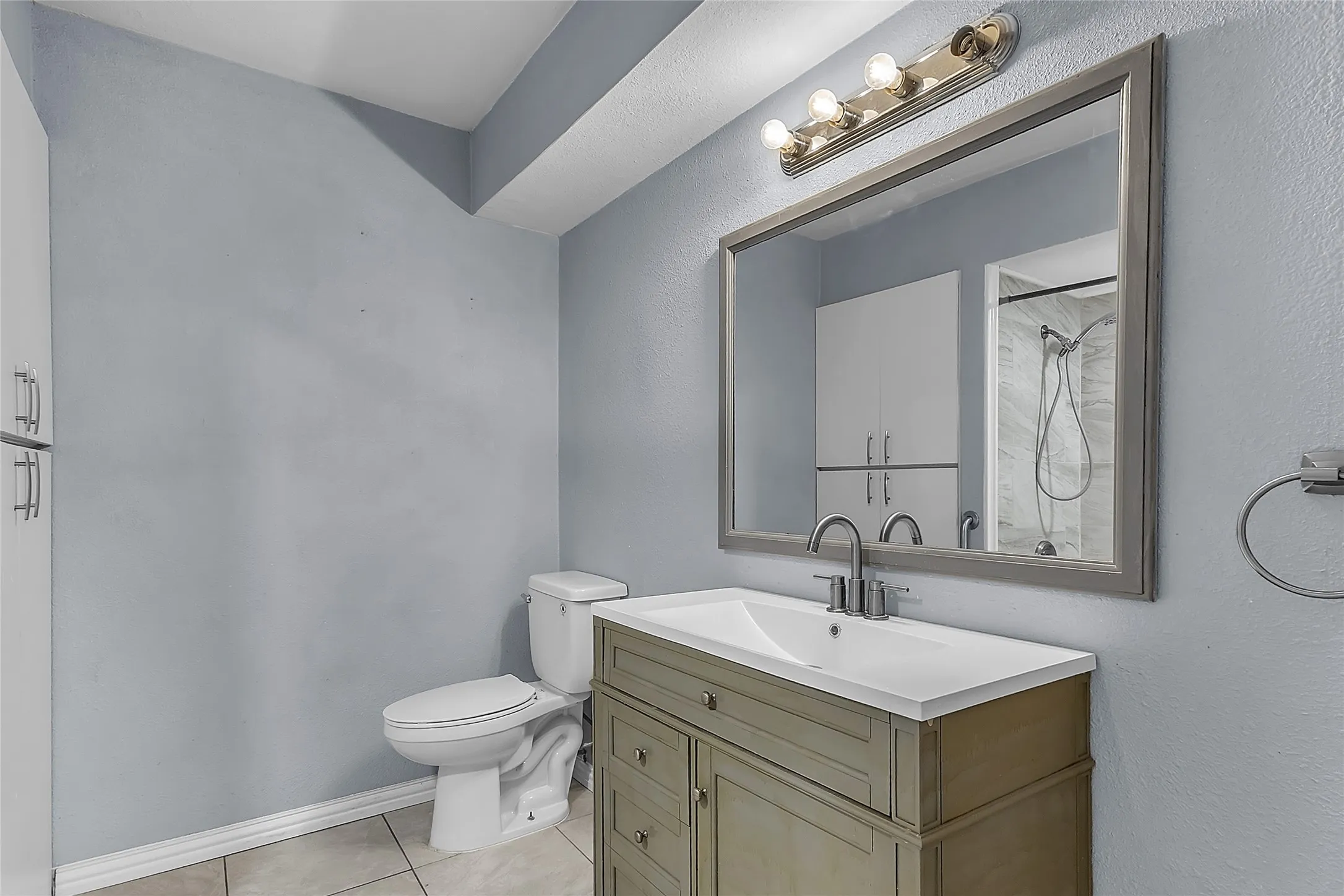 Bathroom with dual vanity sink, white quartz countertop, framed mirror, and porcelain tile floors. Natural light from adjacent shower area.