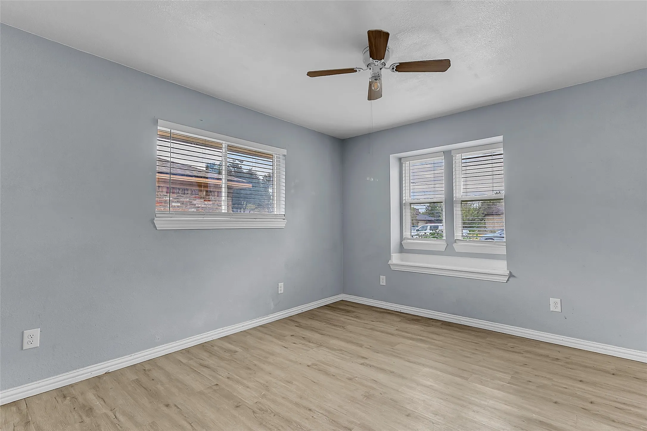 Light-filled bedroom with LVP flooring, ceiling fan, two large windows, and a fresh blue paint finish on the walls.