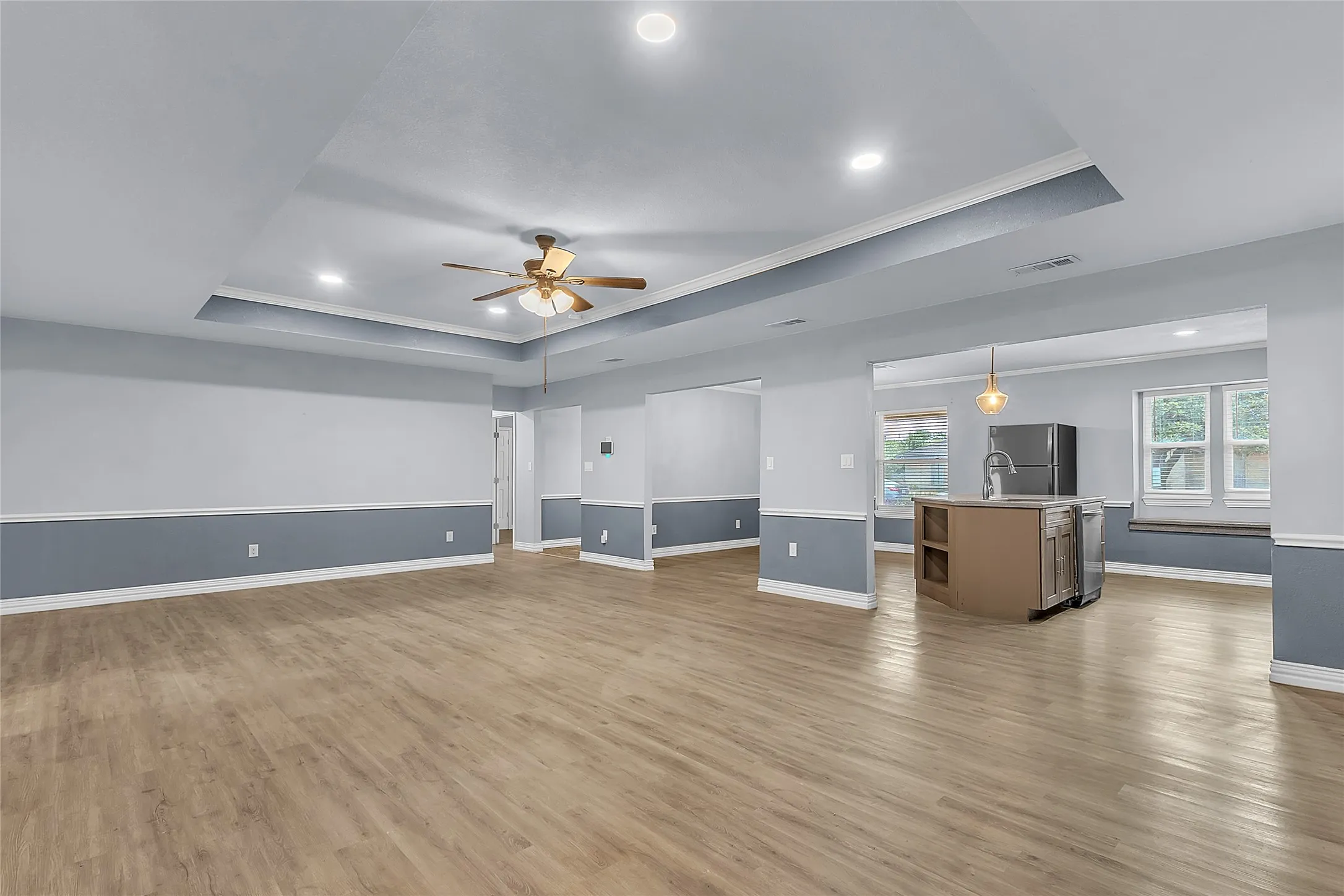 Open-plan living area with hardwood flooring, recessed lighting, and a tray ceiling, leading into a kitchen with stainless steel appliances.