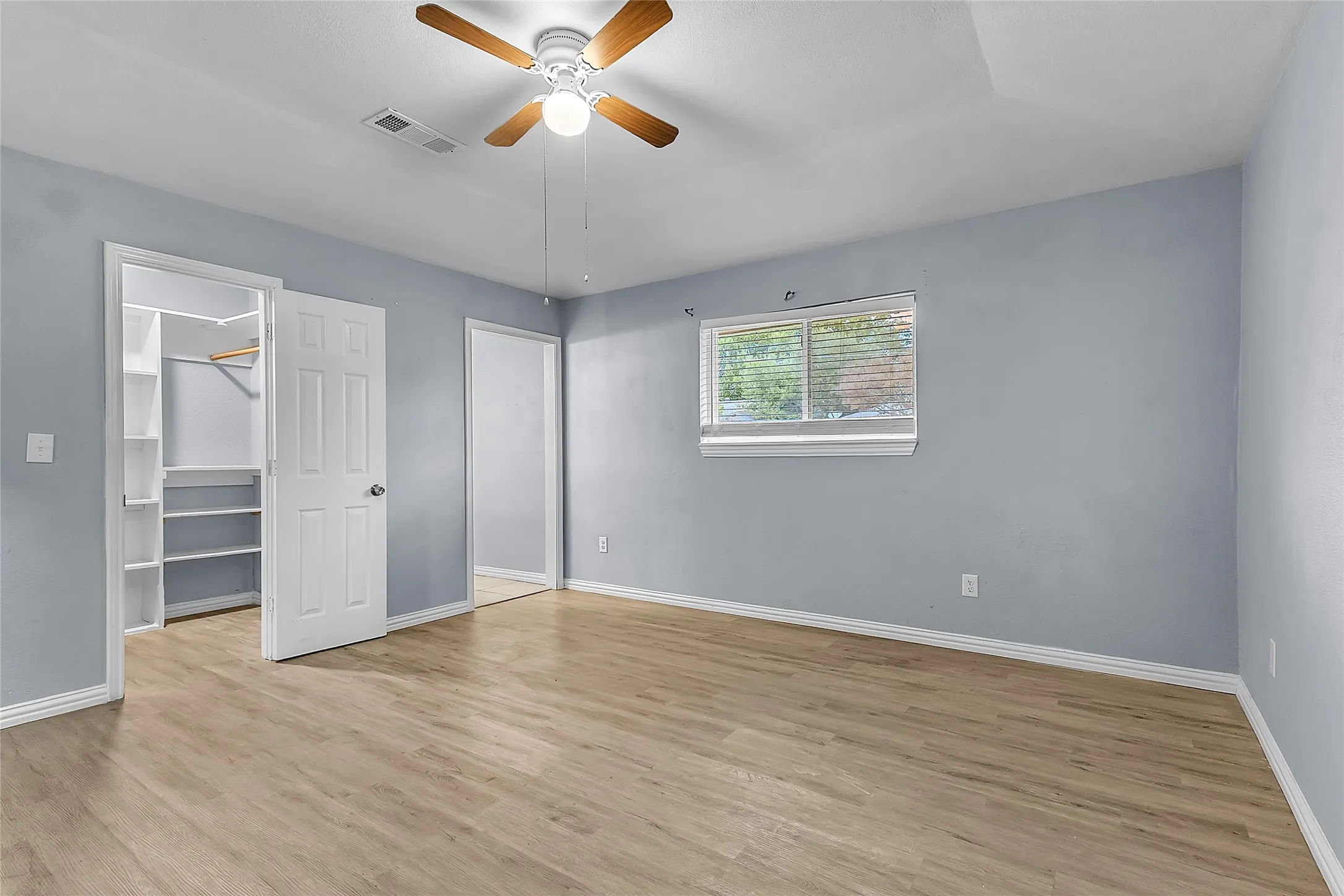 Bedroom with light-colored walls, wood-like flooring, ceiling fan, and a closet with built-in shelving, framed by natural light from a window.
