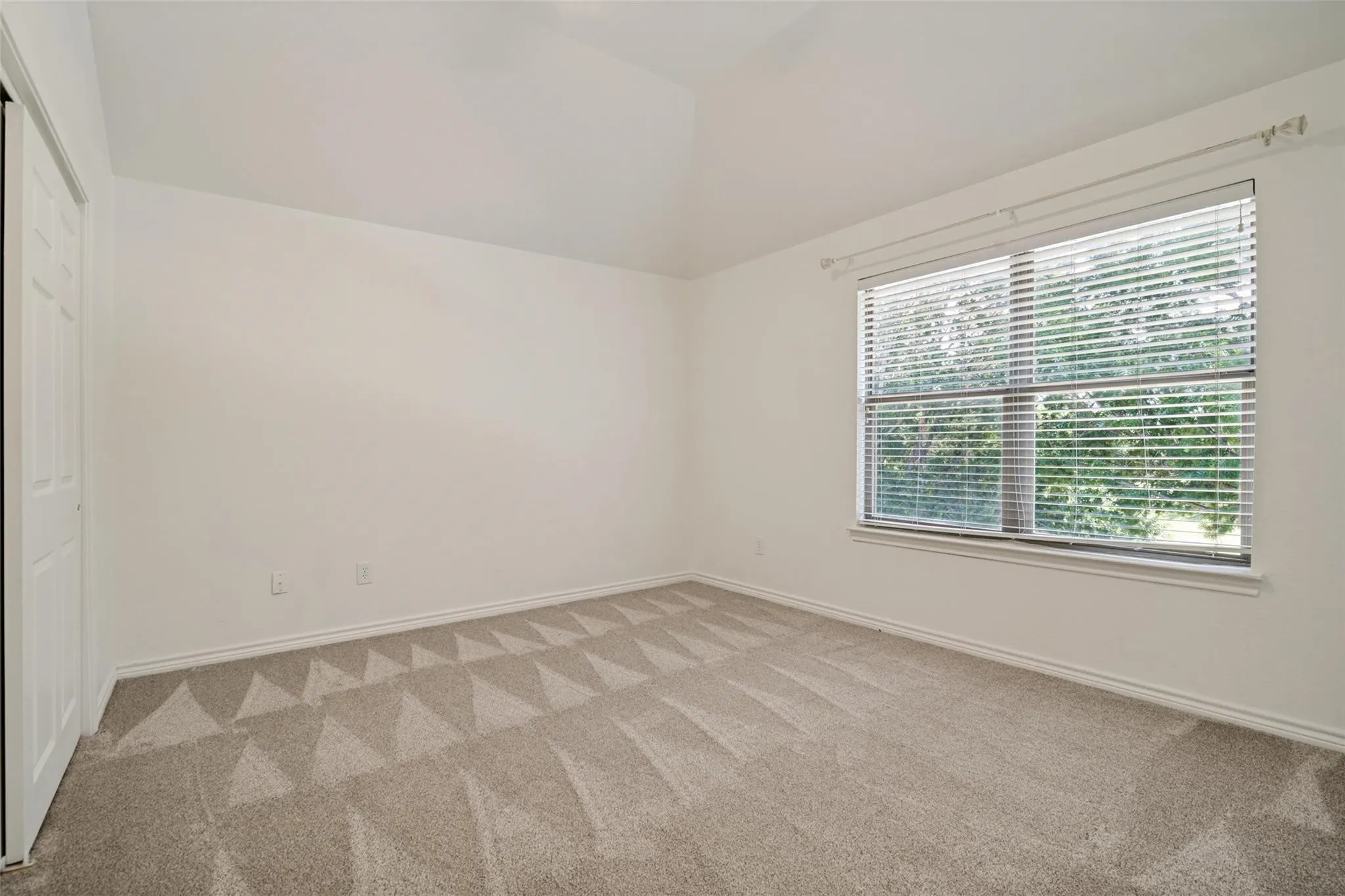 Vaulted ceiling, neutral wall paint, carpeted flooring with geometric pattern, window with blinds, bi-fold closet door