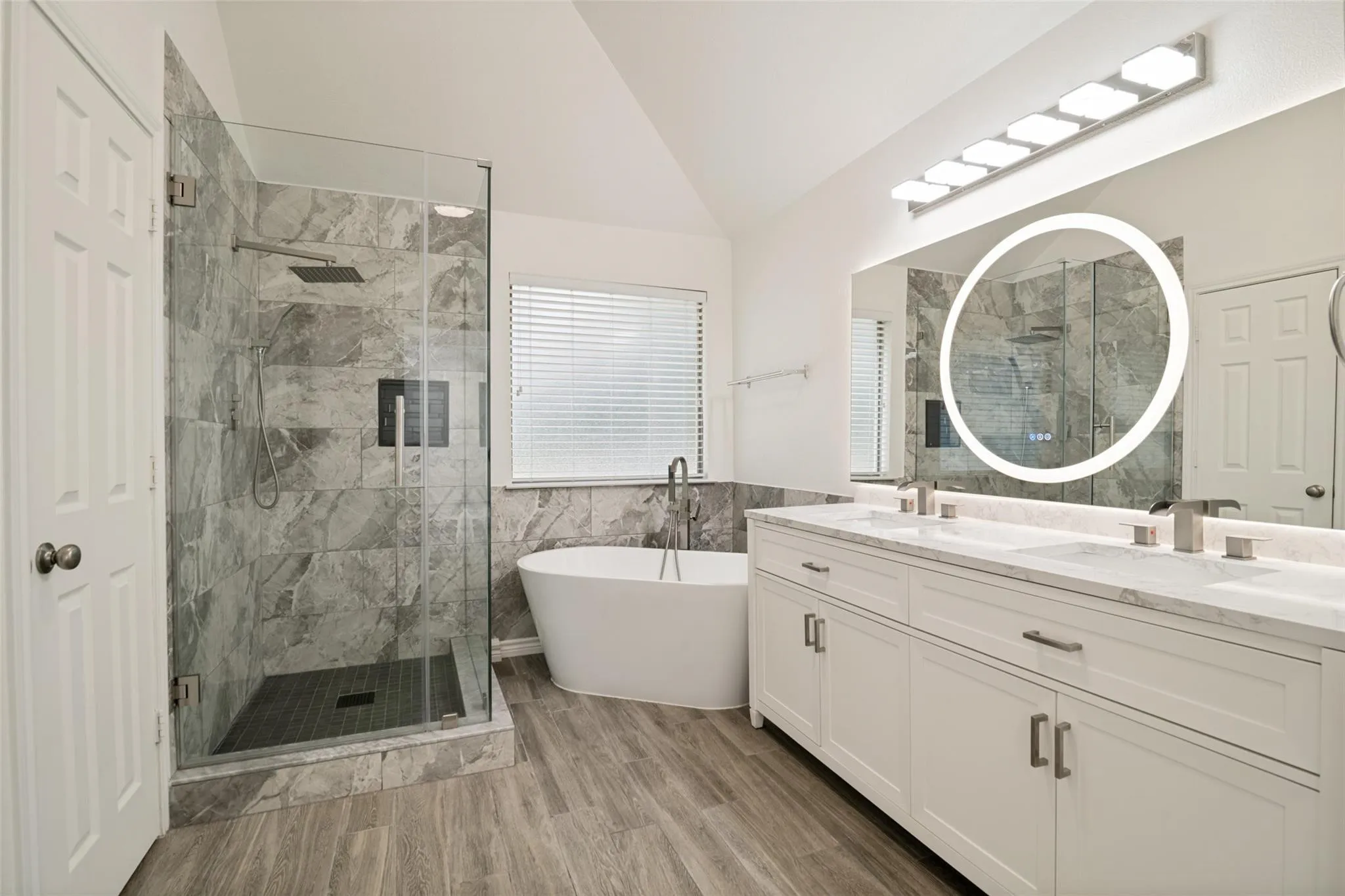 Spacious bathroom featuring a frameless glass shower enclosure, freestanding soaking tub, dual vanity with undermount sinks, and wood-finish flooring