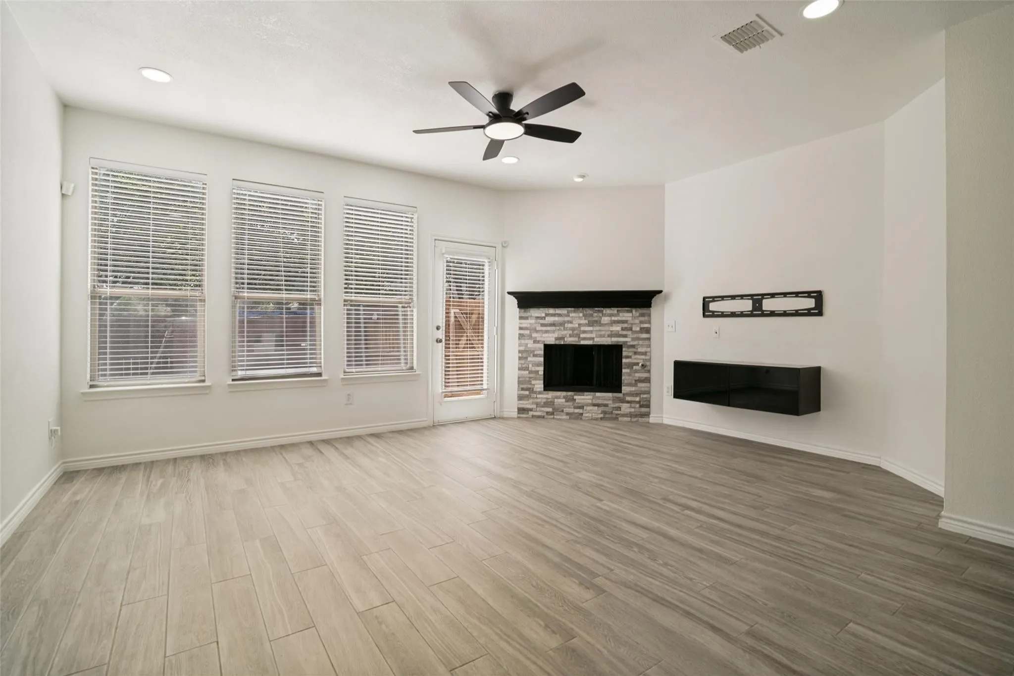 Spacious living area featuring wood-finish flooring, a stacked stone fireplace with a dark mantel, multiple windows, a ceiling fan, and recessed lighting