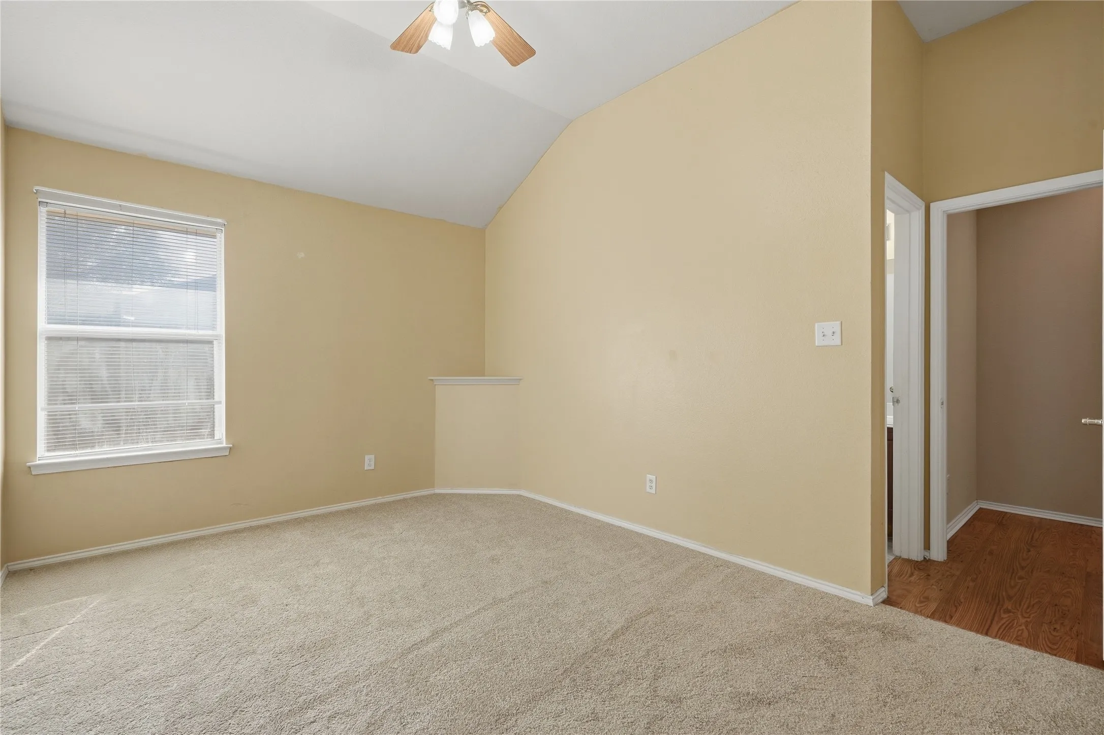 Vaulted ceiling with an integrated ceiling fan, light beige carpeting, a large window, and a doorway with wood-finish flooring