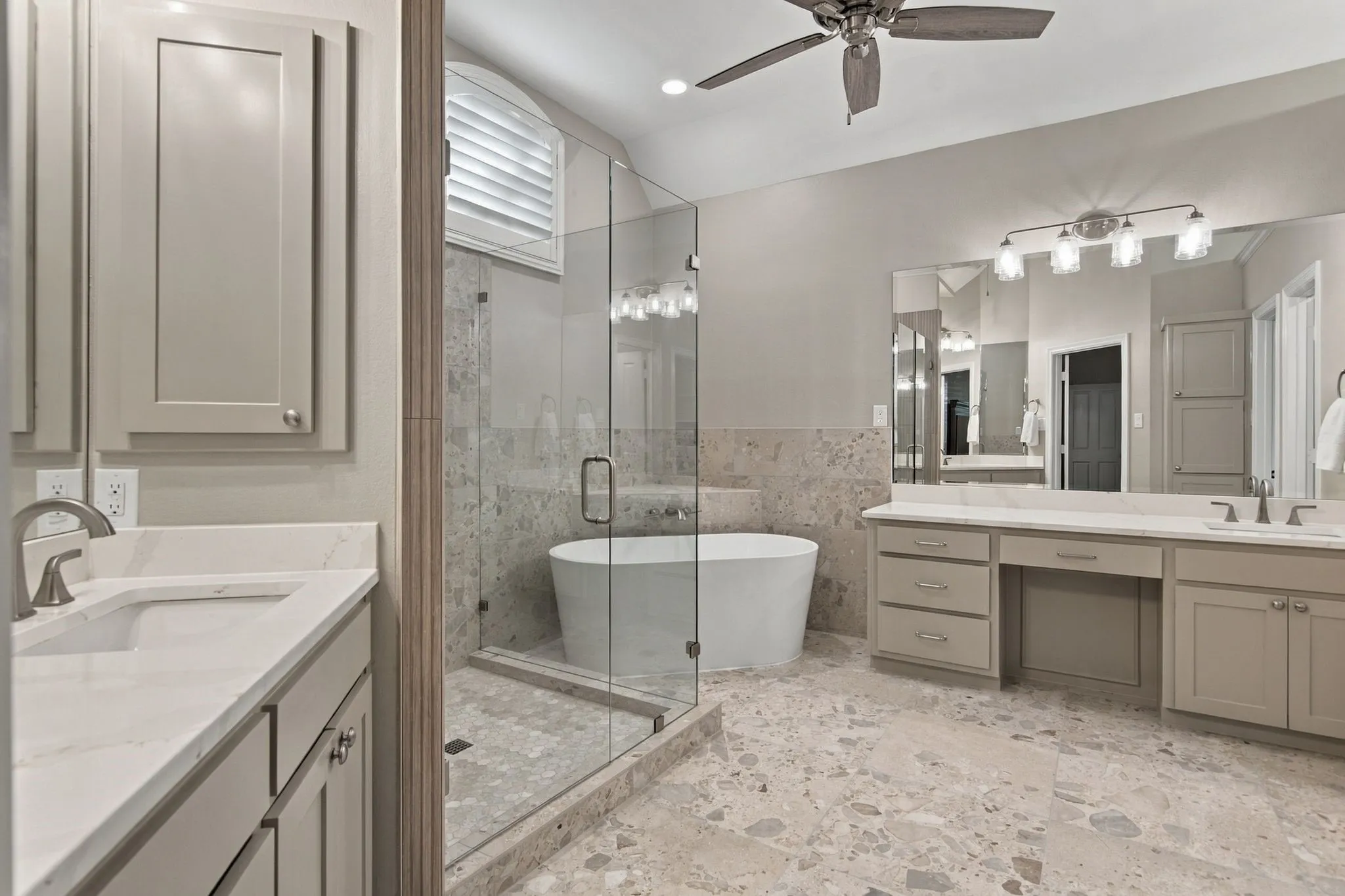 Completely new primary bath with dual vanities, quartz countertops, custom cabinets, designer fixtures,  freestanding soaker bathtub, glass-enclosed shower, ceiling fan, and stone flooring.