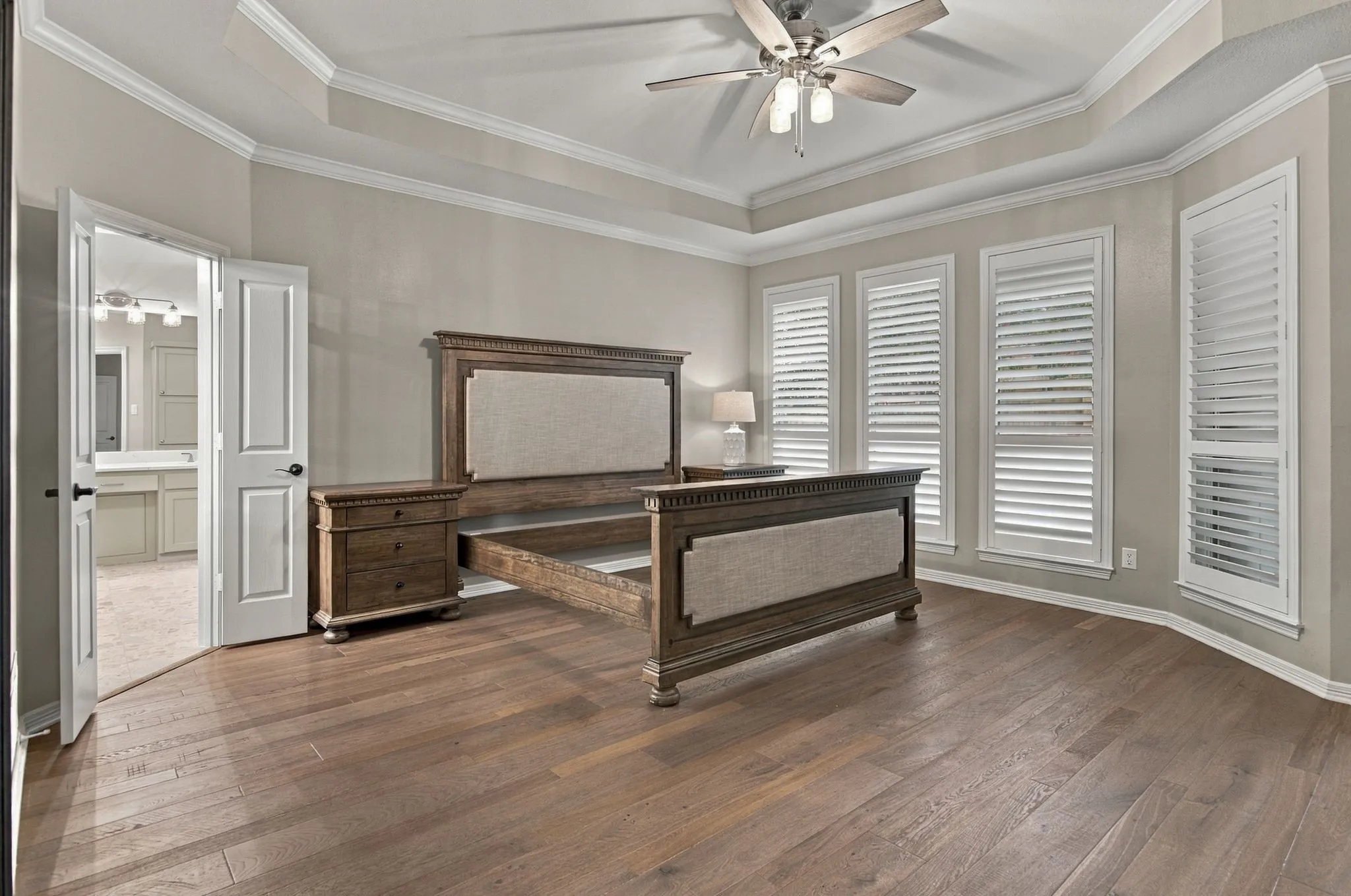 Oversized primary suite featuring tray ceiling with crown molding, wood flooring, ceiling fan, and multiple windows with plantation shutters.