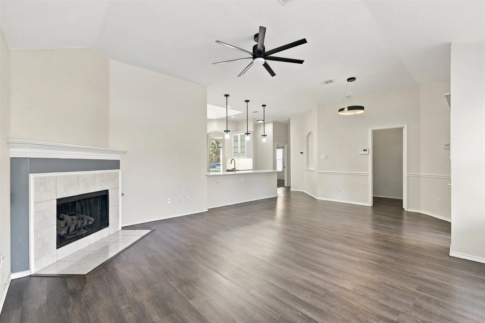 Vaulted ceiling with a black ceiling fan, wood-finish flooring, fireplace with a tile surround and mantel, contemporary pendant lighting, and wainscoting