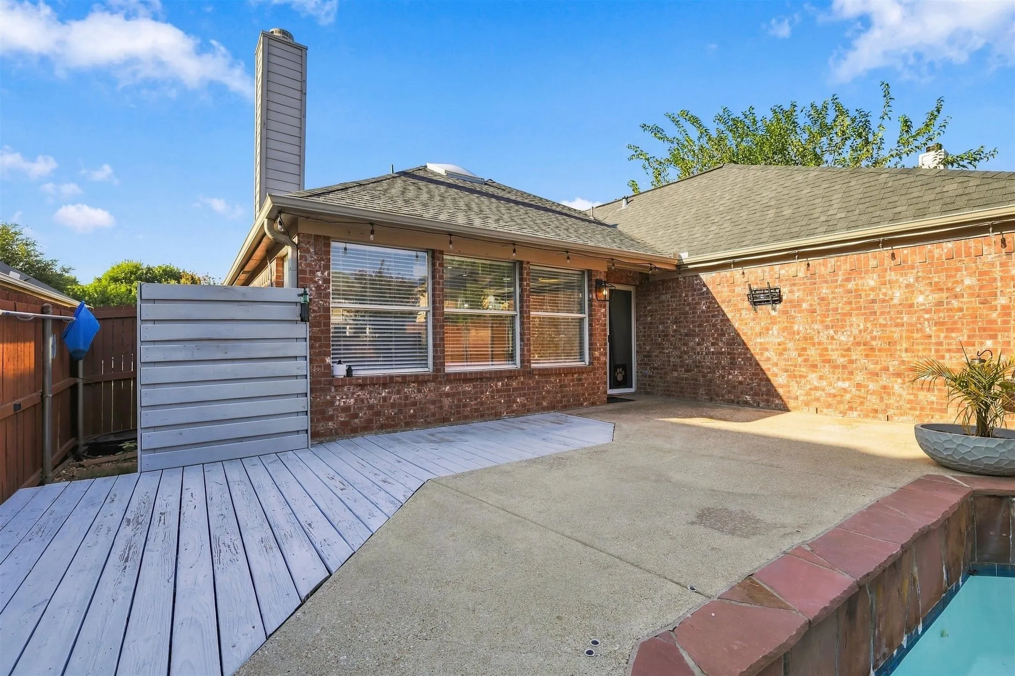 Brick exterior with a prominent chimney, shingled roof, and a combination of concrete patio and wood-finish decking