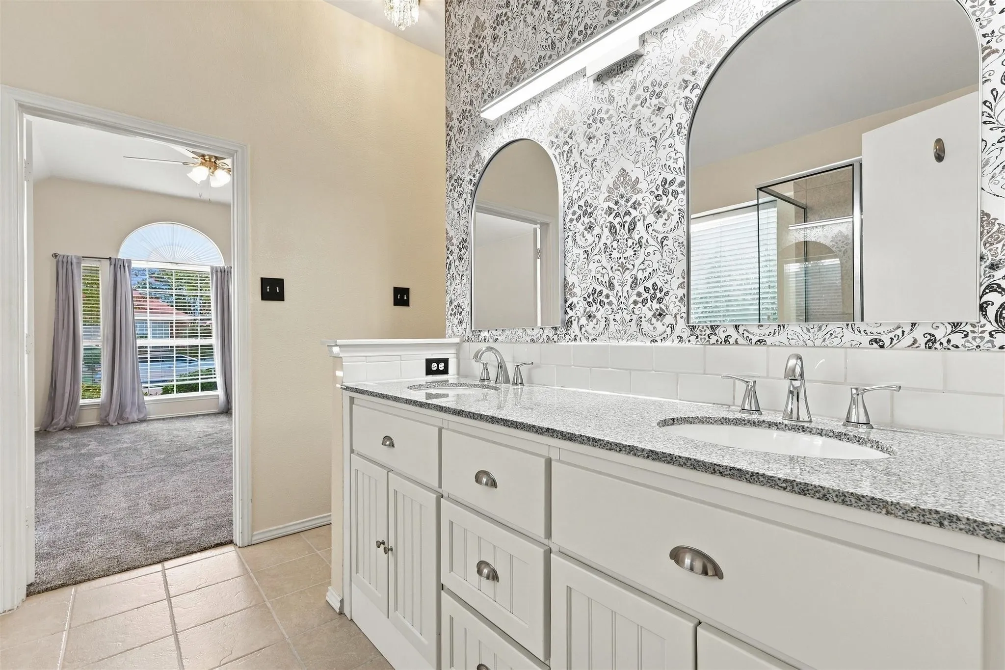 Dual arched mirrors with patterned wallcovering, white subway tile backsplash, dual undermount sinks with chrome fixtures, and a white vanity with paneled doors and drawers