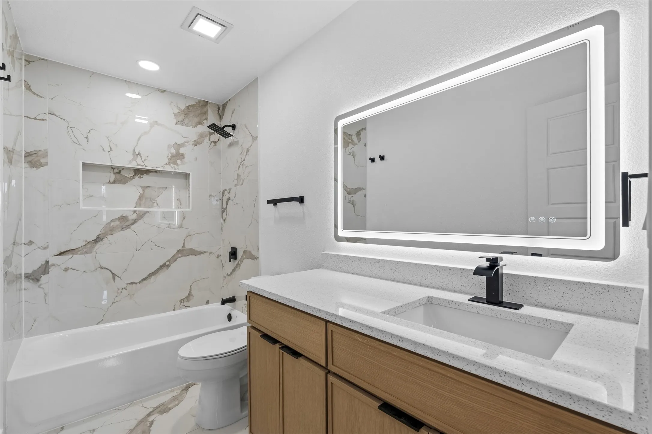 Tiled shower with a niche, tub, wood-finish vanity, integrated sink, and an illuminated mirror
