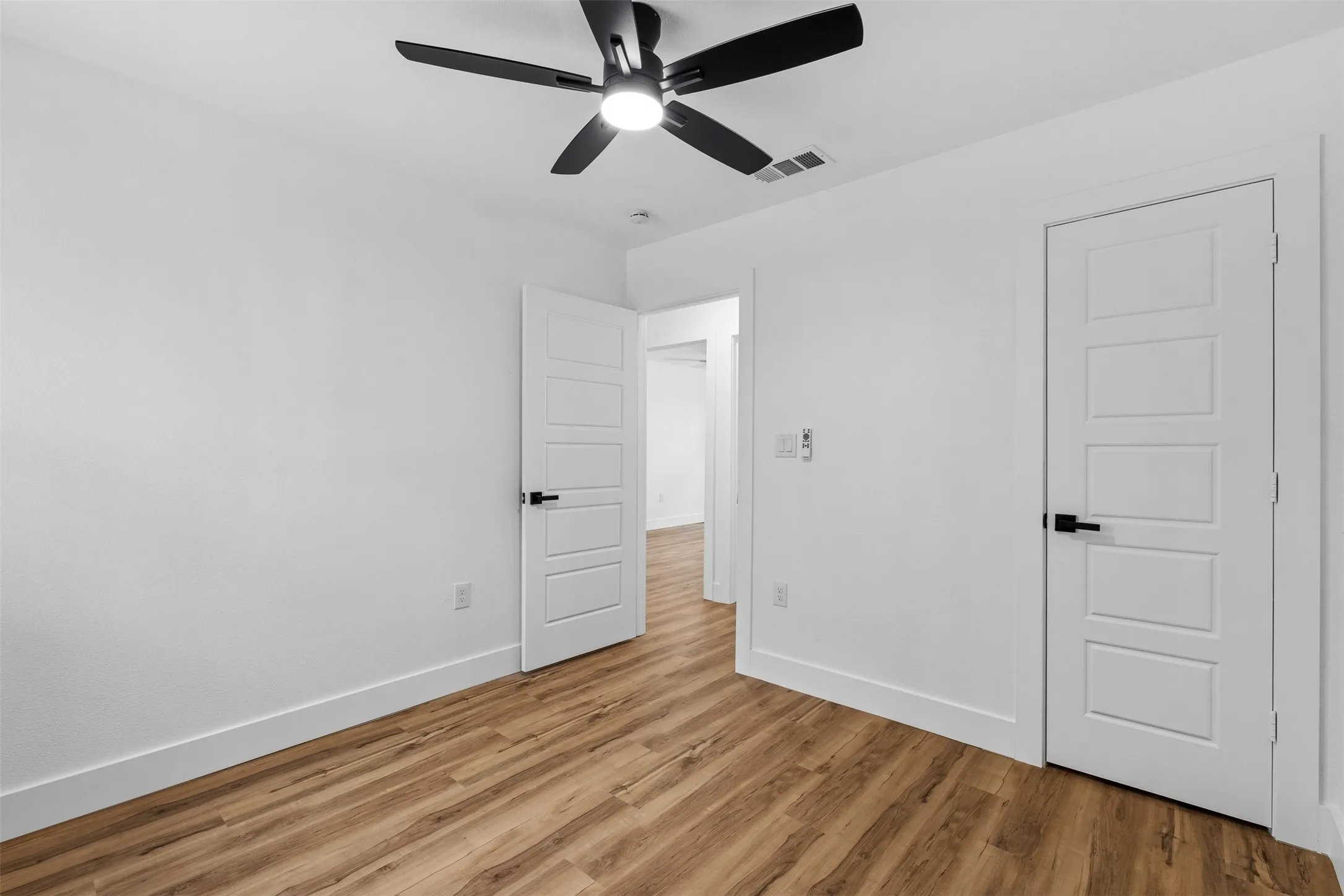 Wood-finish flooring, white paneled doors with black hardware, and a ceiling fan with integrated lighting