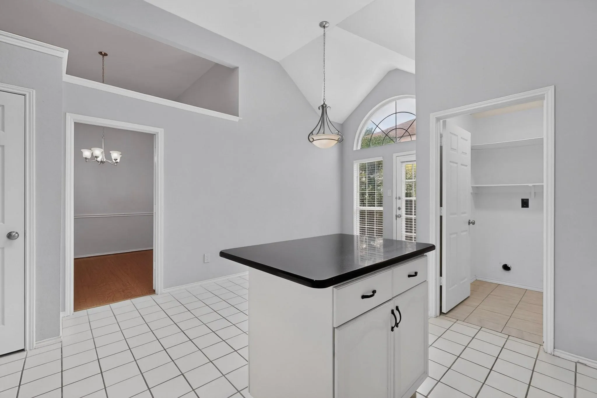 Vaulted ceiling with an arched window, white tile flooring, white cabinetry island with black countertop, and an exterior door with integrated blinds