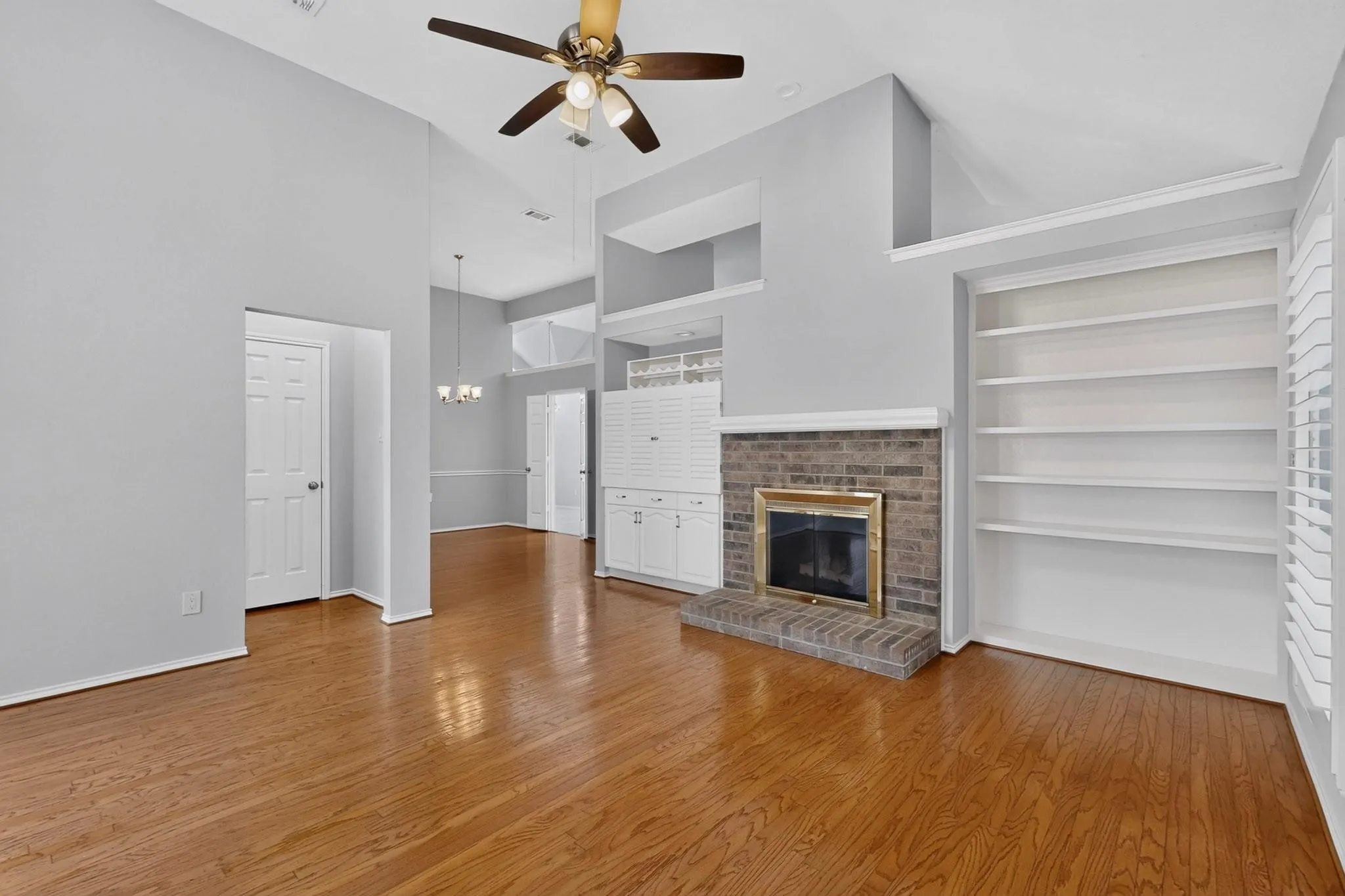 Vaulted ceiling with a ceiling fan, wood-finish flooring, brick fireplace with a brass-tone insert, built-in shelving, and built-in white cabinetry