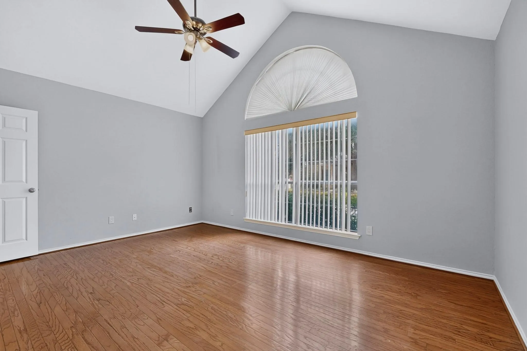 Vaulted ceiling with a ceiling fan, arched transom window, large window with vertical blinds, wood-finish flooring, and a paneled door