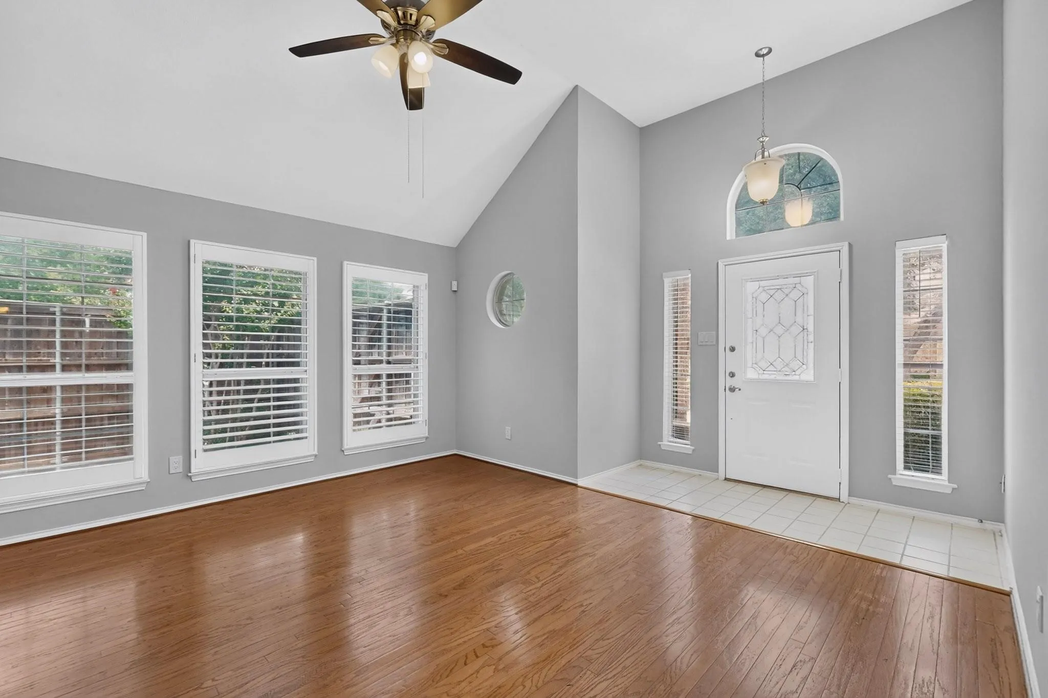 Vaulted ceiling with a ceiling fan, wood-finish flooring, multiple windows with plantation shutters, white entry door with decorative glass, and a tiled entry area
