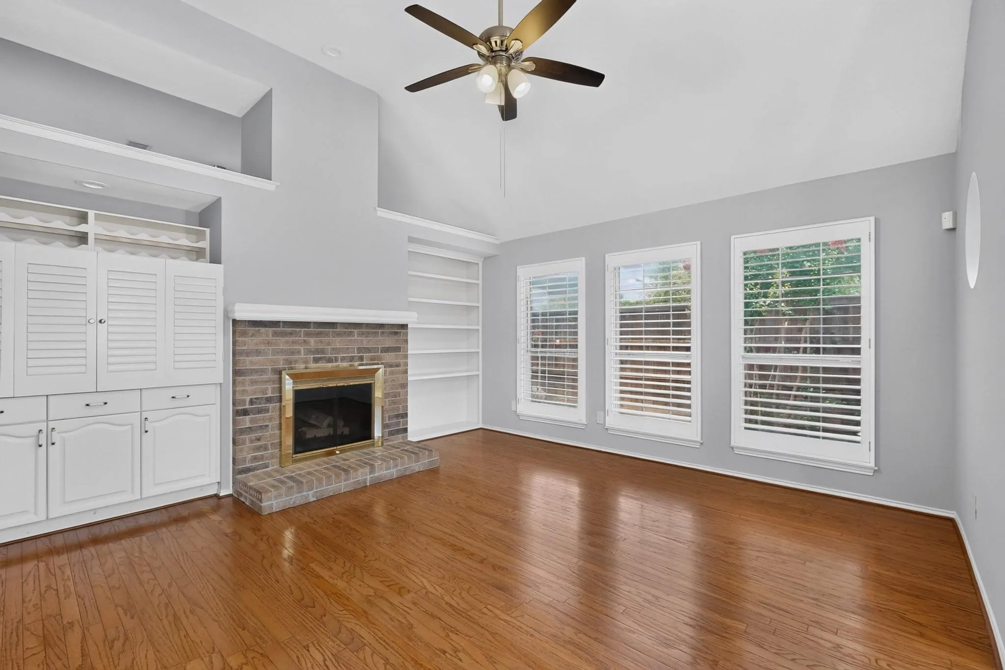 Spacious great room featuring warm wood-finish flooring, a brick fireplace with built-in surround, white shutter cabinetry with integrated storage, multiple glass doors with integrated blinds flooding the space with natural light, and a brass ceiling fan
