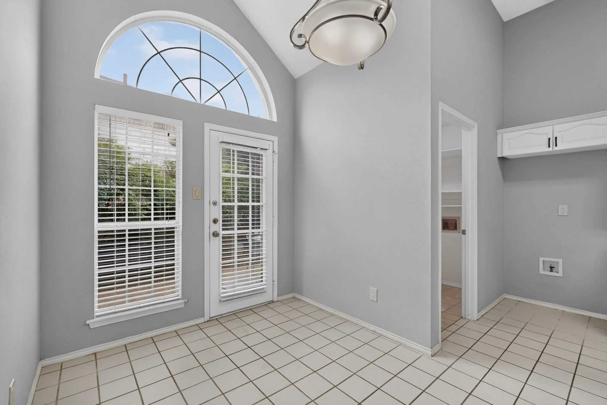 Ceramic tile flooring, white shaker-style wall cabinets, arched transom window, glass-panel exterior door, and recessed shelving