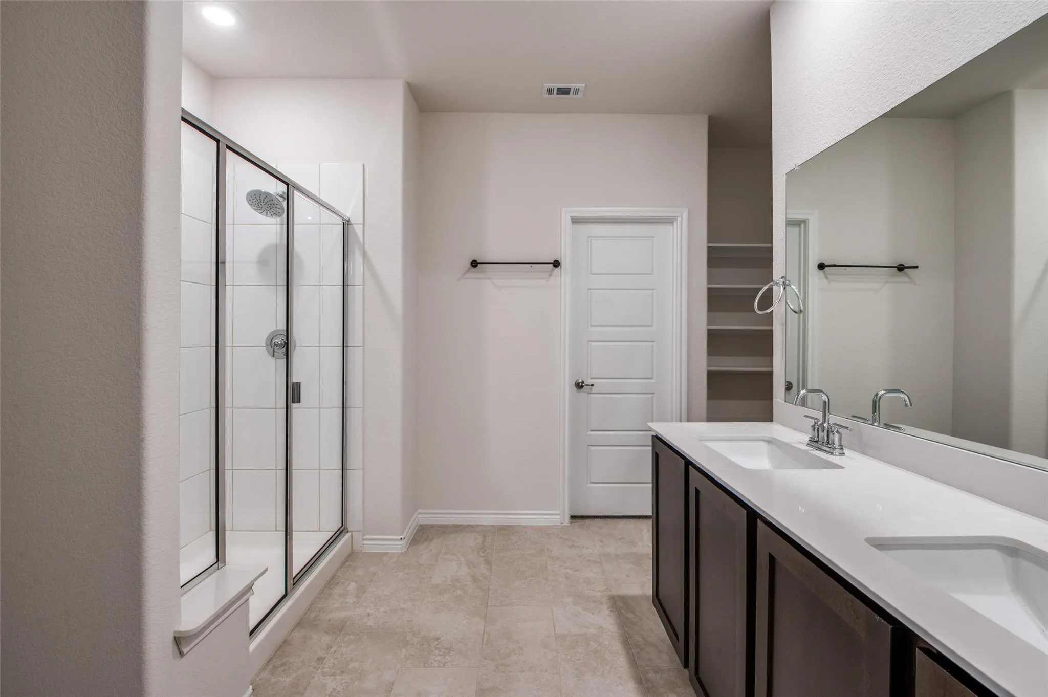 Spacious bathroom featuring a dual vanity with white countertops, a large framed mirror, a walk-in shower with clear glass enclosure, and tiled flooring