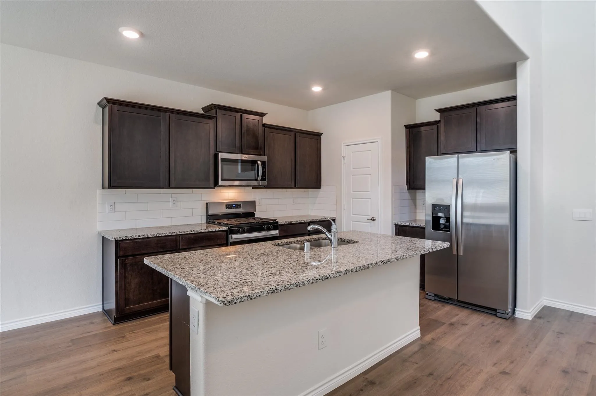 Kitchen featuring an island with a stainless steel sink, granite countertops, dark wood cabinetry, and wood-finish flooring