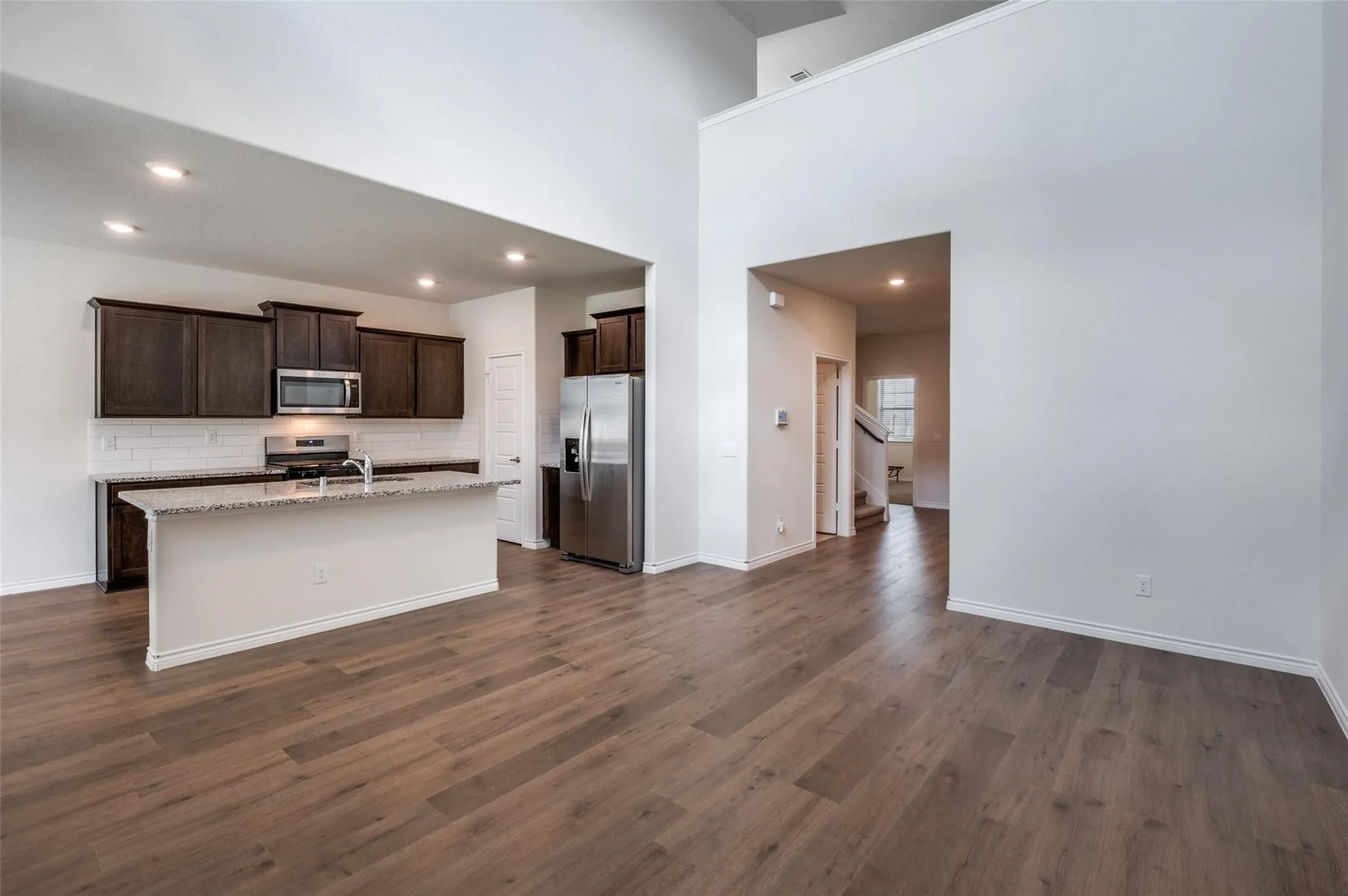 Open-concept living space with wood-finish flooring, high ceilings, recessed lighting, and a kitchen featuring dark wood cabinetry, stainless steel appliances, and a granite-topped island with a sink