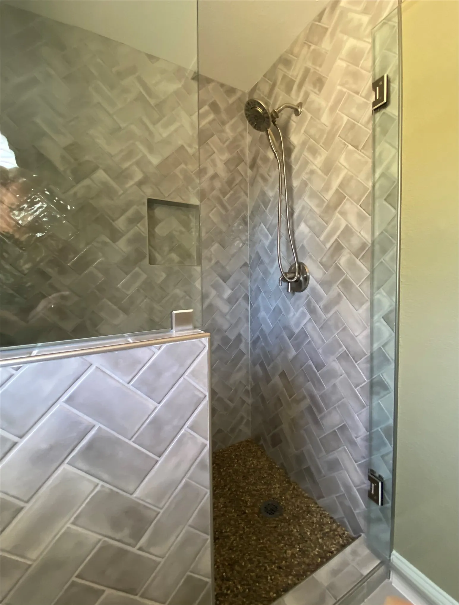 Glass-enclosed shower featuring herringbone tile walls, a built-in shower niche, a dual shower head system, and a pebble-finish floor