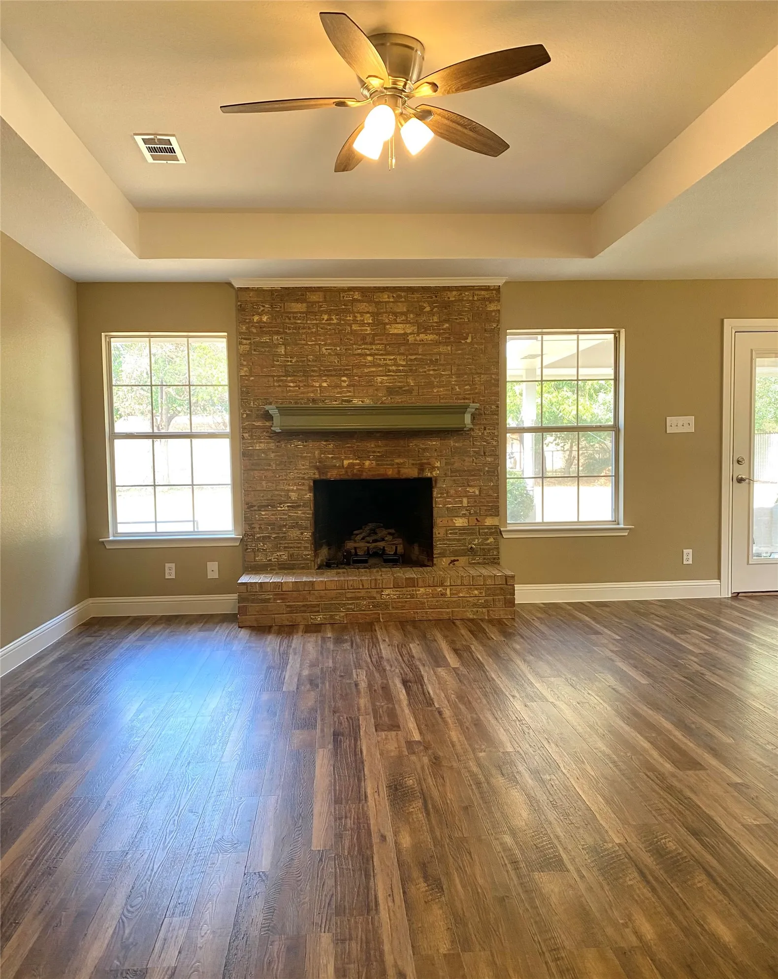 Wood-finish flooring, brick fireplace with a mantel, tray ceiling, and a ceiling fan with integrated lighting
