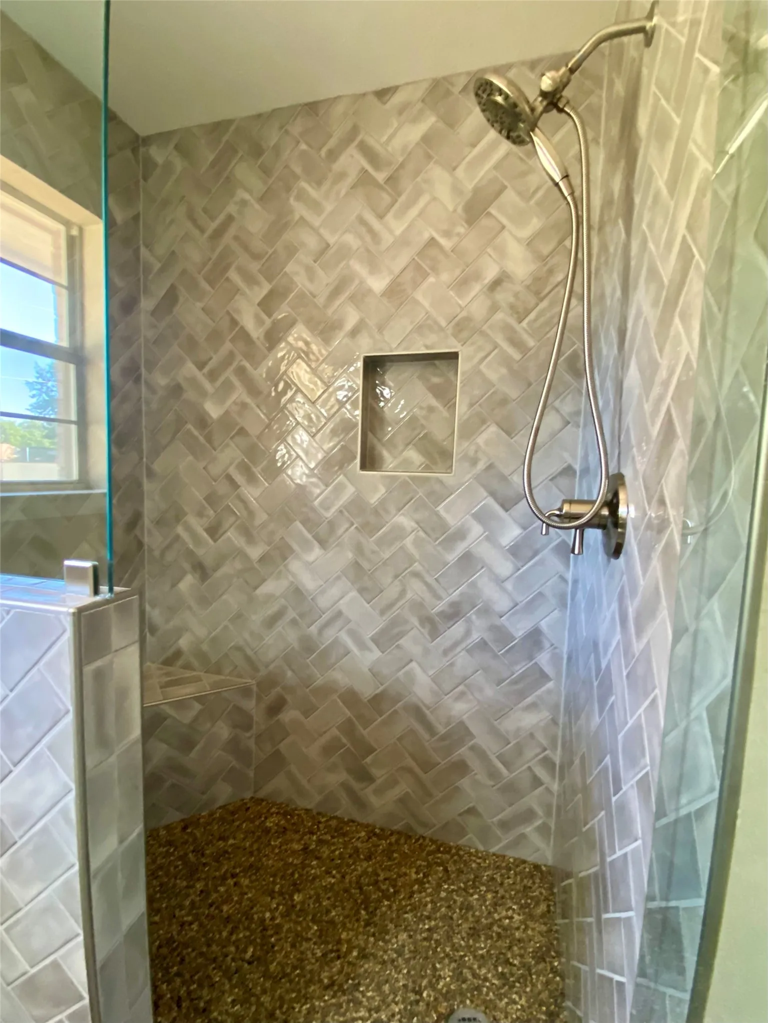 Shower with a natural pebble floor, herringbone-patterned tile walls, built-in recessed shelf, built-in tiled bench, and a multi-function showerhead