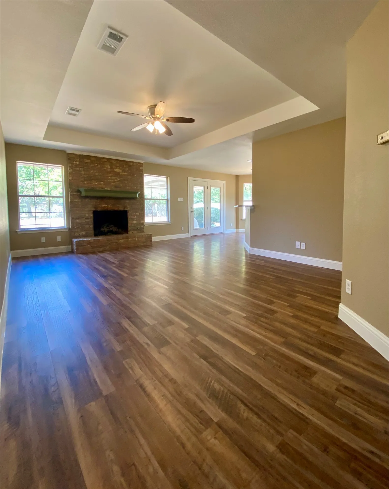 Wood-finish flooring throughout, brick fireplace with mantel, tray ceiling with ceiling fan, dual windows, and French doors