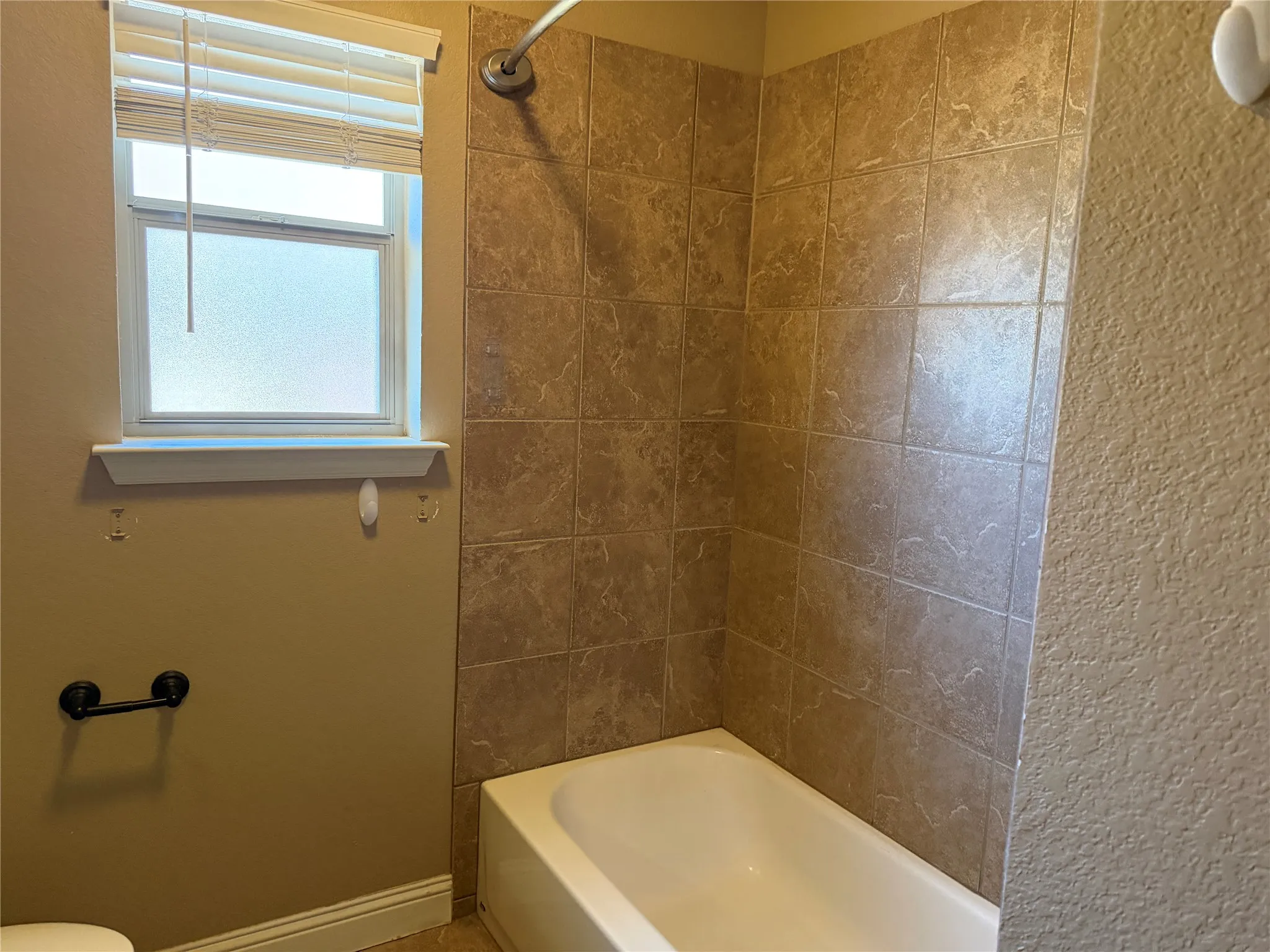 Tub and shower combination featuring a tiled surround and a window with blinds