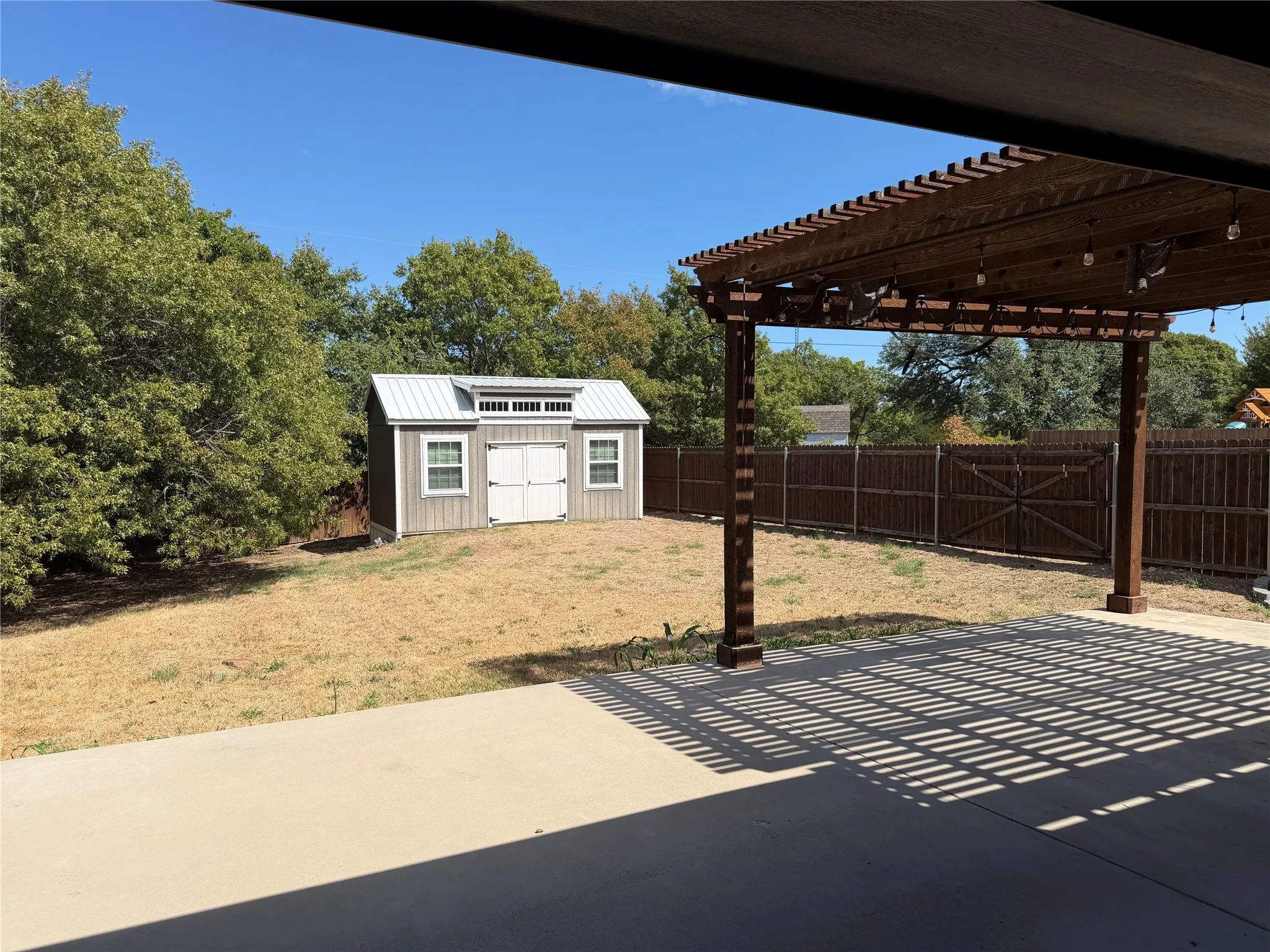 Expansive backyard featuring a stained wood pergola, concrete patio, privacy fencing, and a storage shed with double doors and windows