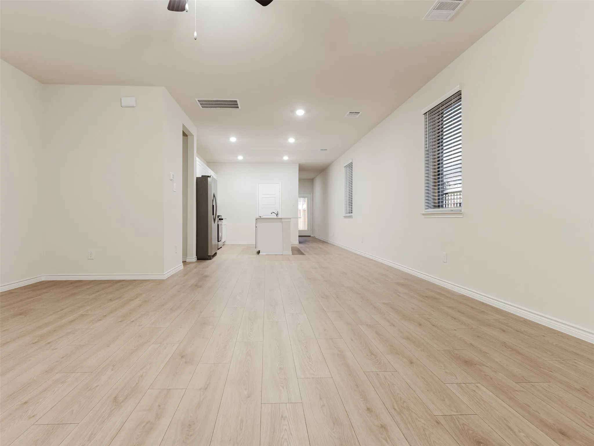 Spacious open-concept interior featuring light wood-finish flooring, recessed lighting, and a stainless steel refrigerator