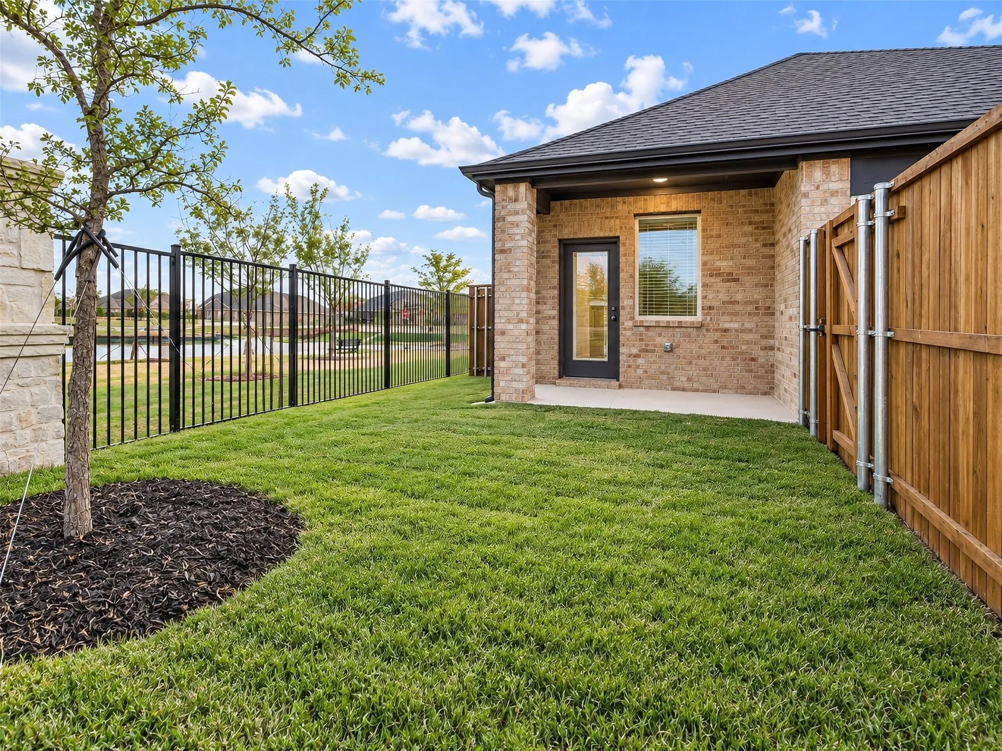 Expansive lawn with a black metal fence, a wooden privacy fence, a brick exterior, and a covered concrete patio