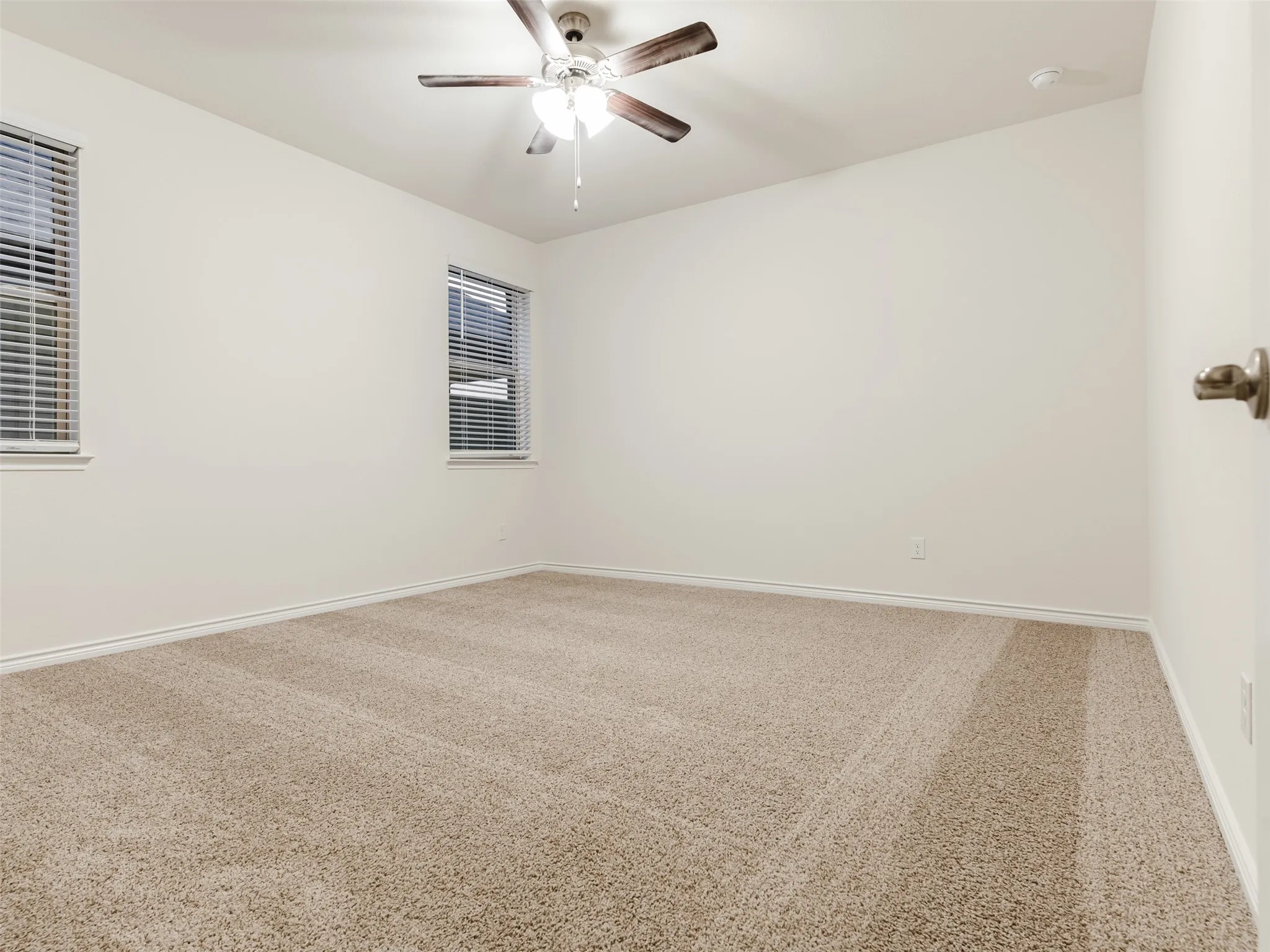 Neutral wall paint, plush carpeting, two windows, and a ceiling fan with light fixture