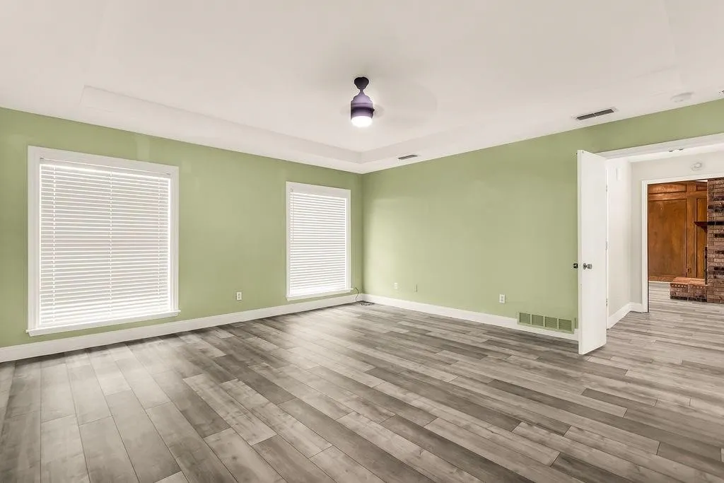 Spacious room featuring wood-finish flooring, two windows with blinds, a tray ceiling, and an overhead light fixture